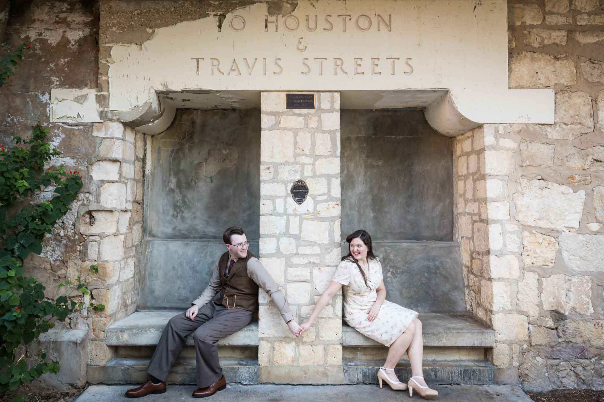 Couple holding hands while sitting on stone benches separated by stone wall during a River Walk surprise proposal