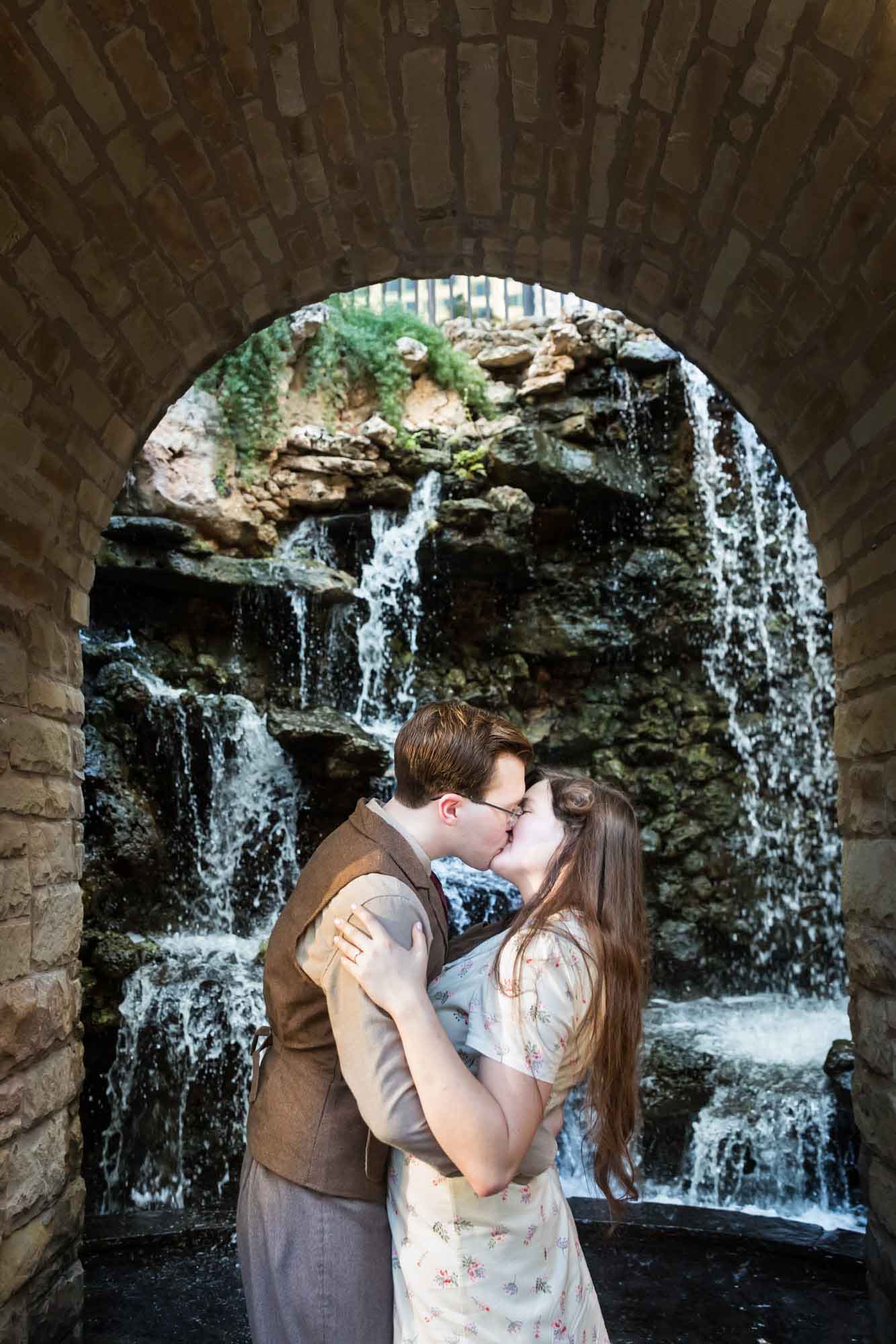 Couple kissing in front of archway in front of waterfall during a River Walk surprise proposal