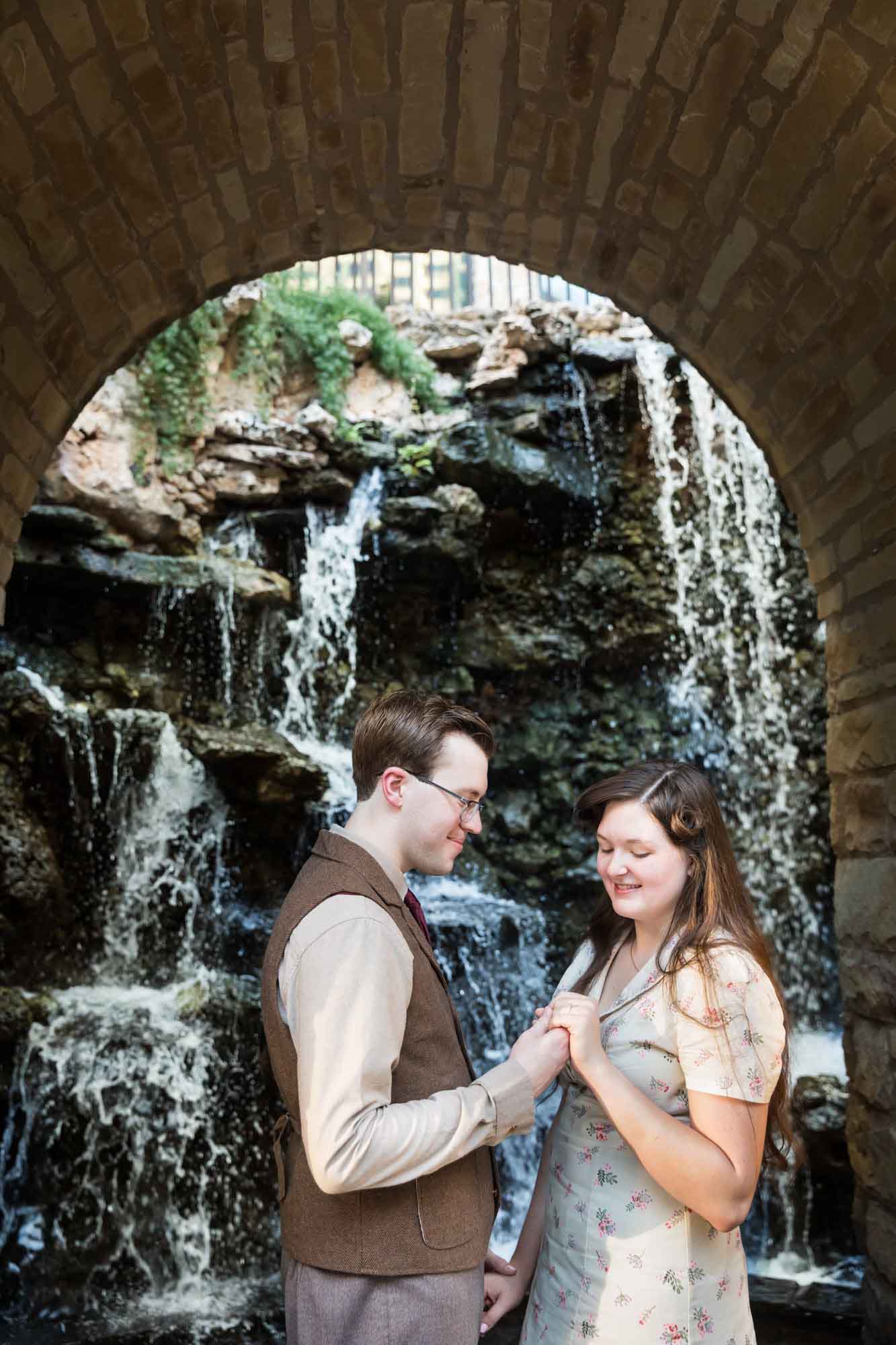 Couple looking at hands in front of archway in front of waterfall during a River Walk surprise proposal