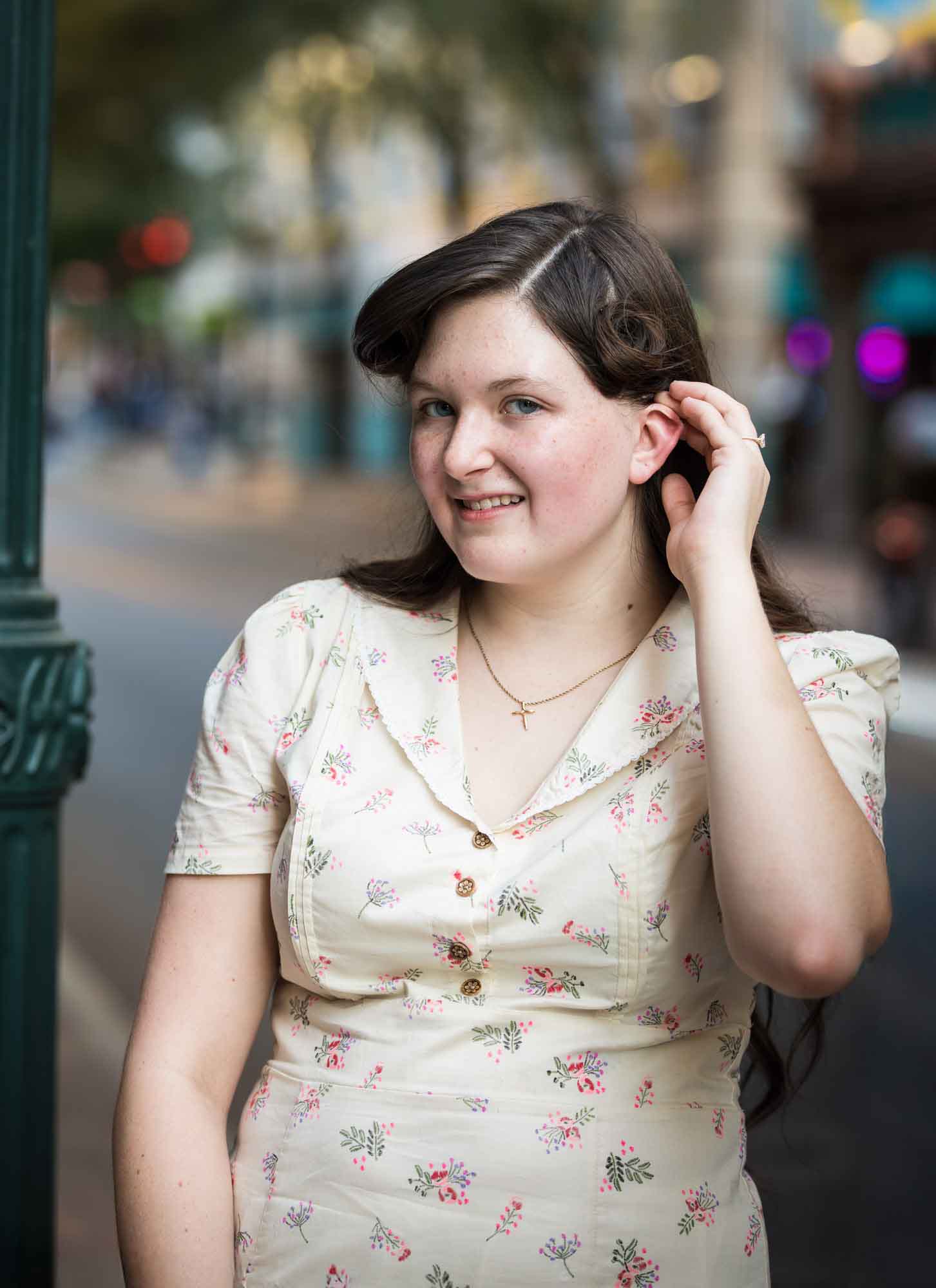 Woman wearing short-sleeved dress pushing hair behind her ear showing engagement ring while standing against green lamp post during a River Walk surprise proposal