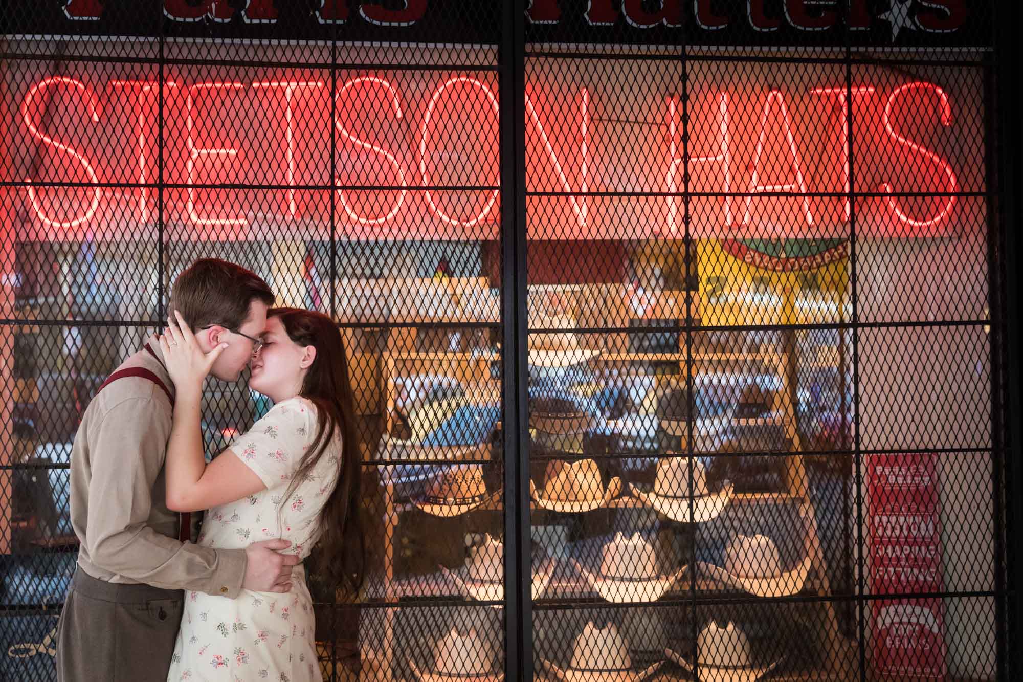 Couple kissing in front of orange neon sign that says 'Stetson Hats' with cowboy hats in window during a River Walk surprise proposal
