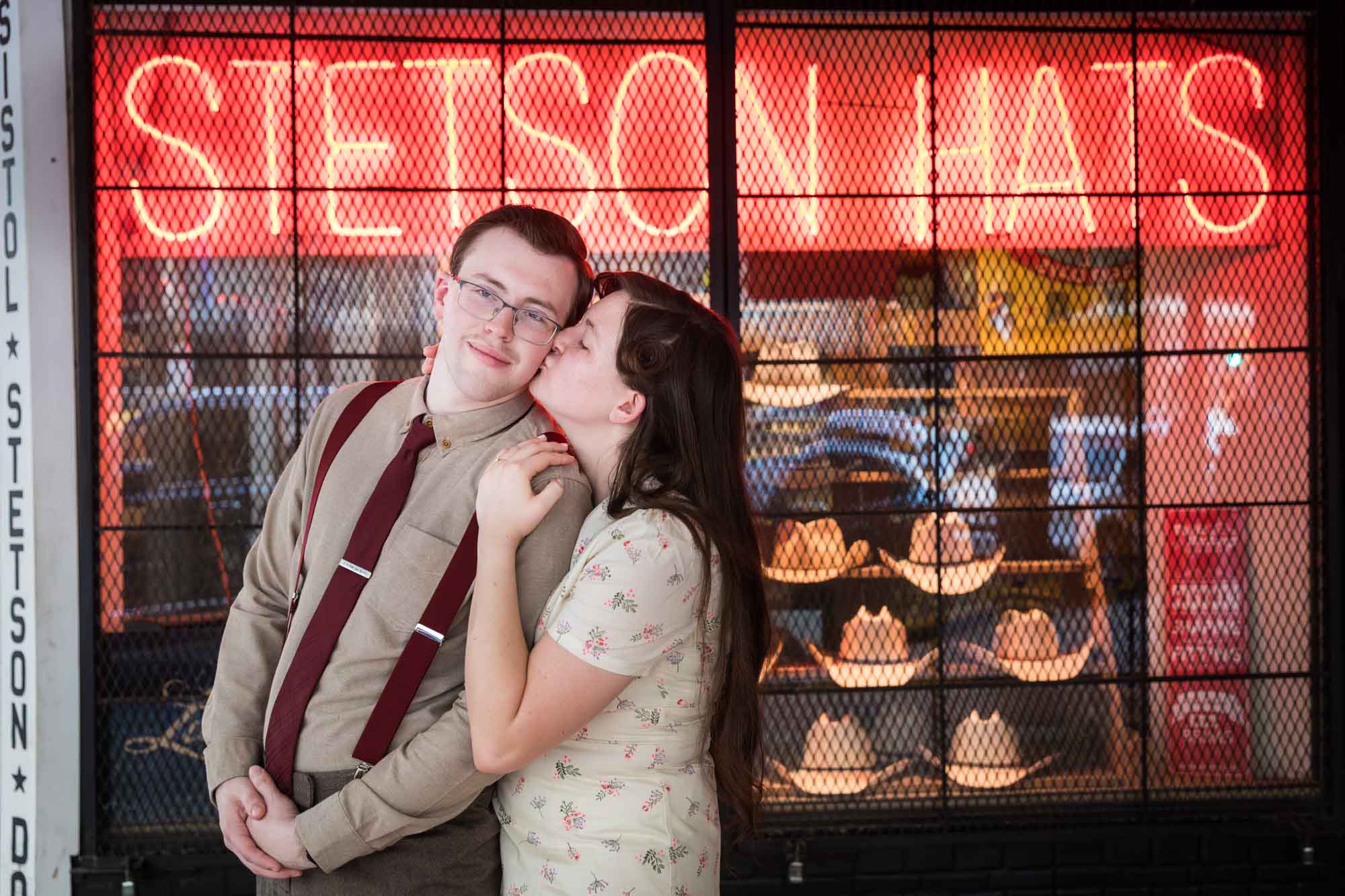 Woman kissing man on the cheek in front of orange neon sign that says 'Stetson Hats' with cowboy hats in window during a River Walk surprise proposal