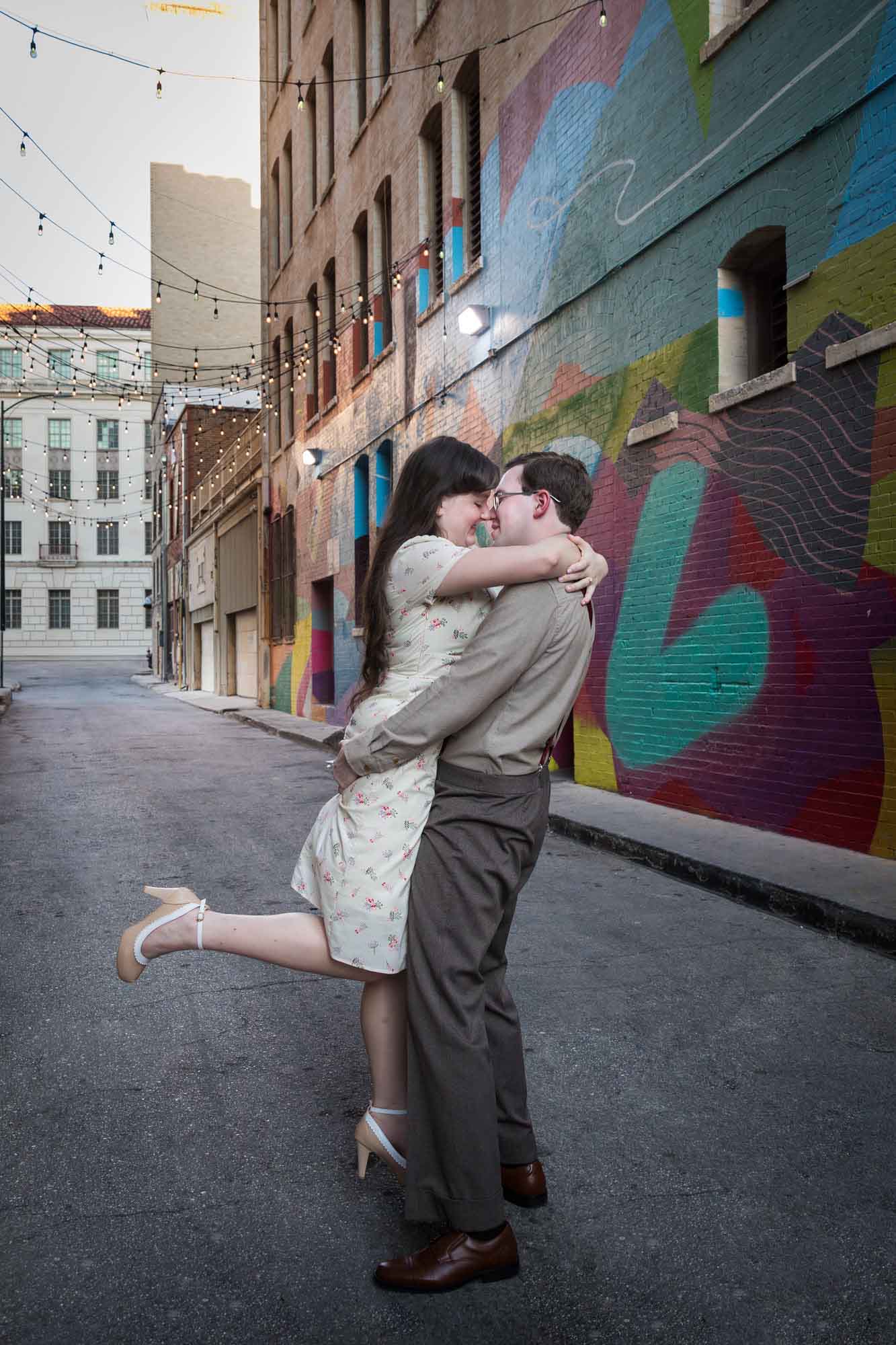 Man lifting woman in the air in front of colored wall murals of Peacock Alley during a River Walk surprise proposal