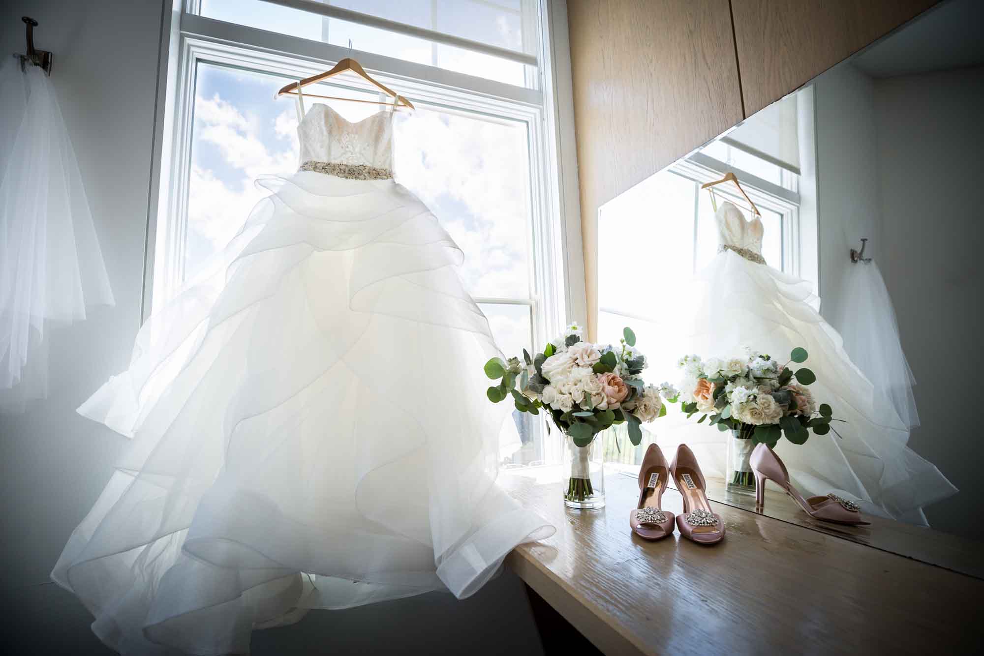 Wedding dress in window reflected in mirror with flower bouquet and pink shows on desk for an article on unexpected wedding photo tips
