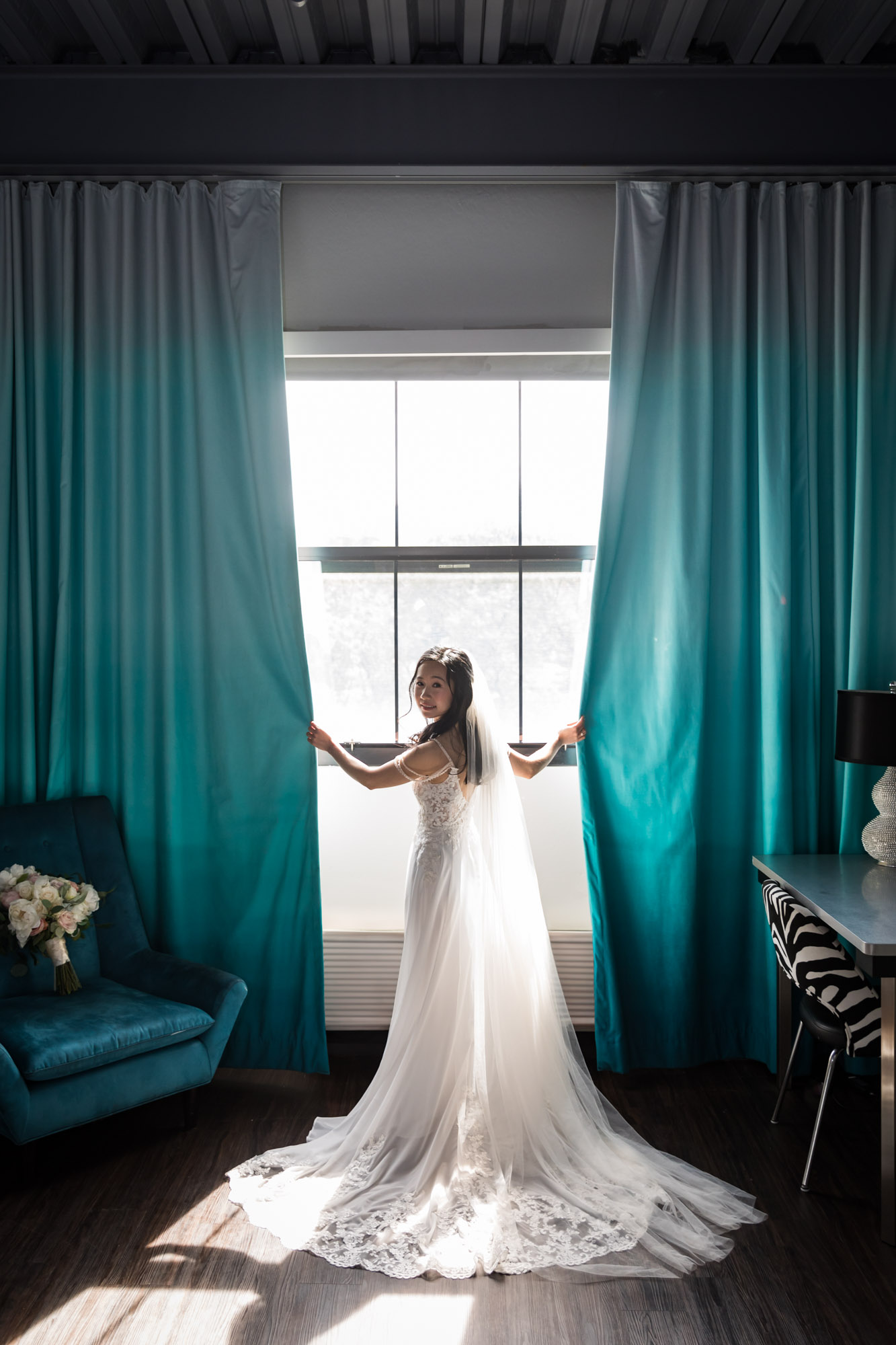 Bride wearing long white dress and veil holding blue curtains in front of window for an article on unexpected wedding photo tips