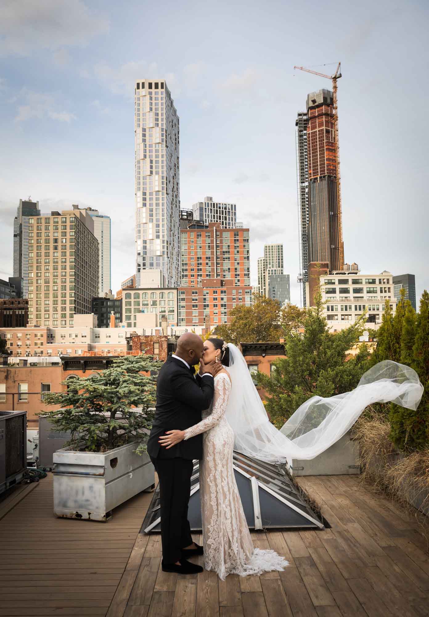Bride with veil extended kissing groom on roof with Brooklyn skyline in background for an article on unexpected wedding photo tips