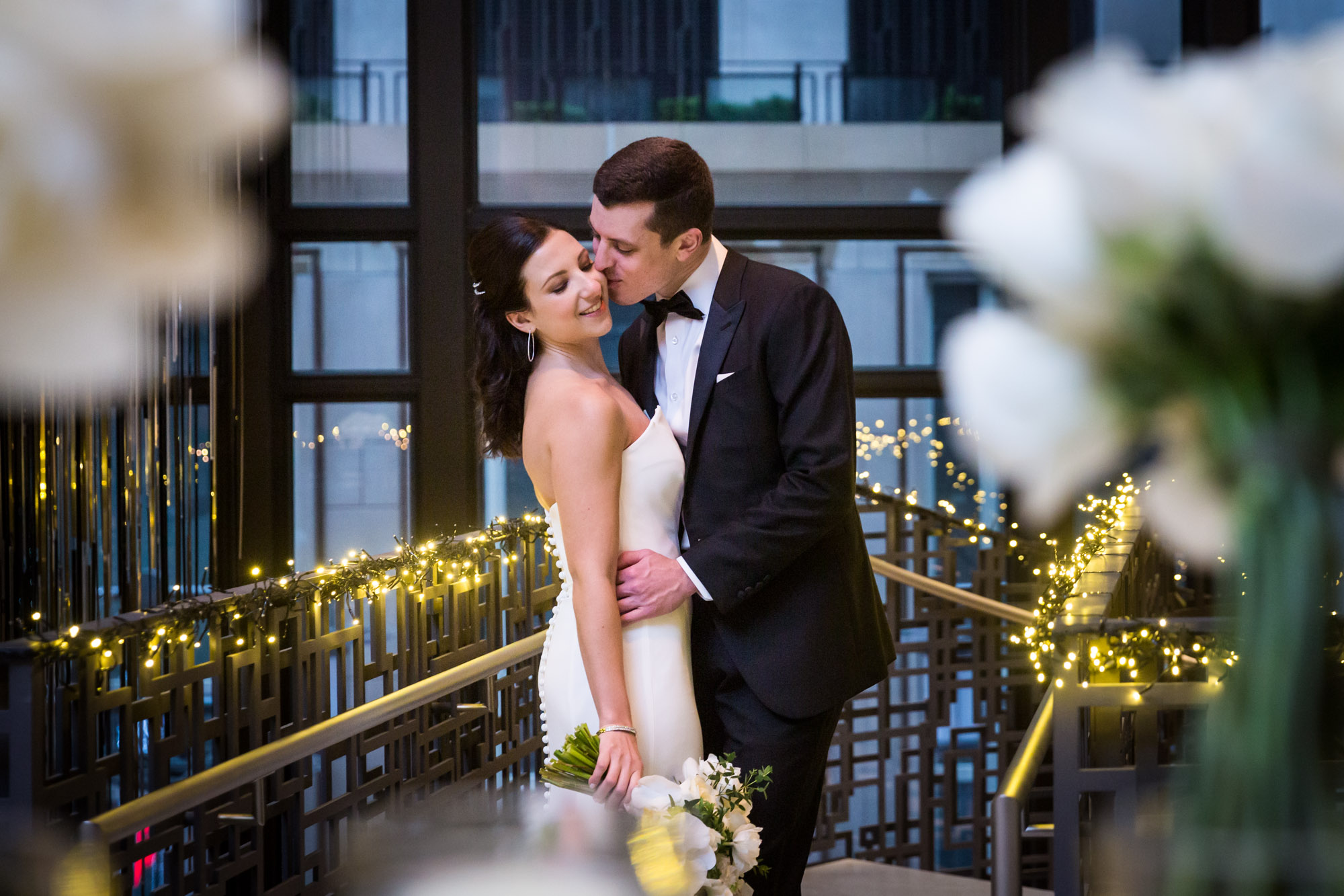 Groom kissing bride on cheek in middle of stair railing wrapped in fairy lights for an article on unexpected wedding photo tips