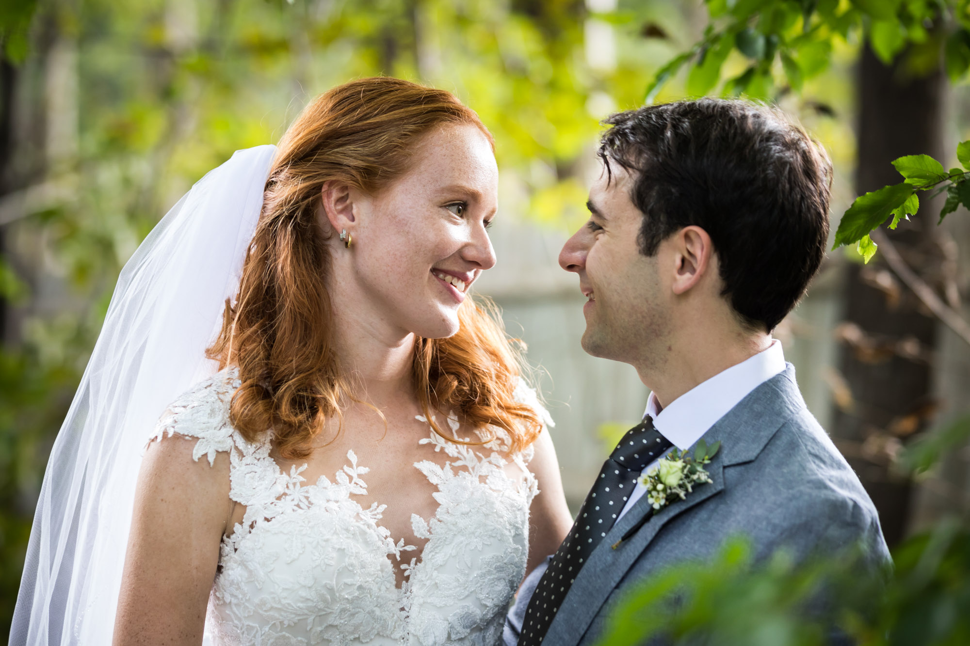 Bride wearing short sleeved dress and veil looking at groom wearing grey blazer in middle of forest for an article on unexpected wedding photo tips