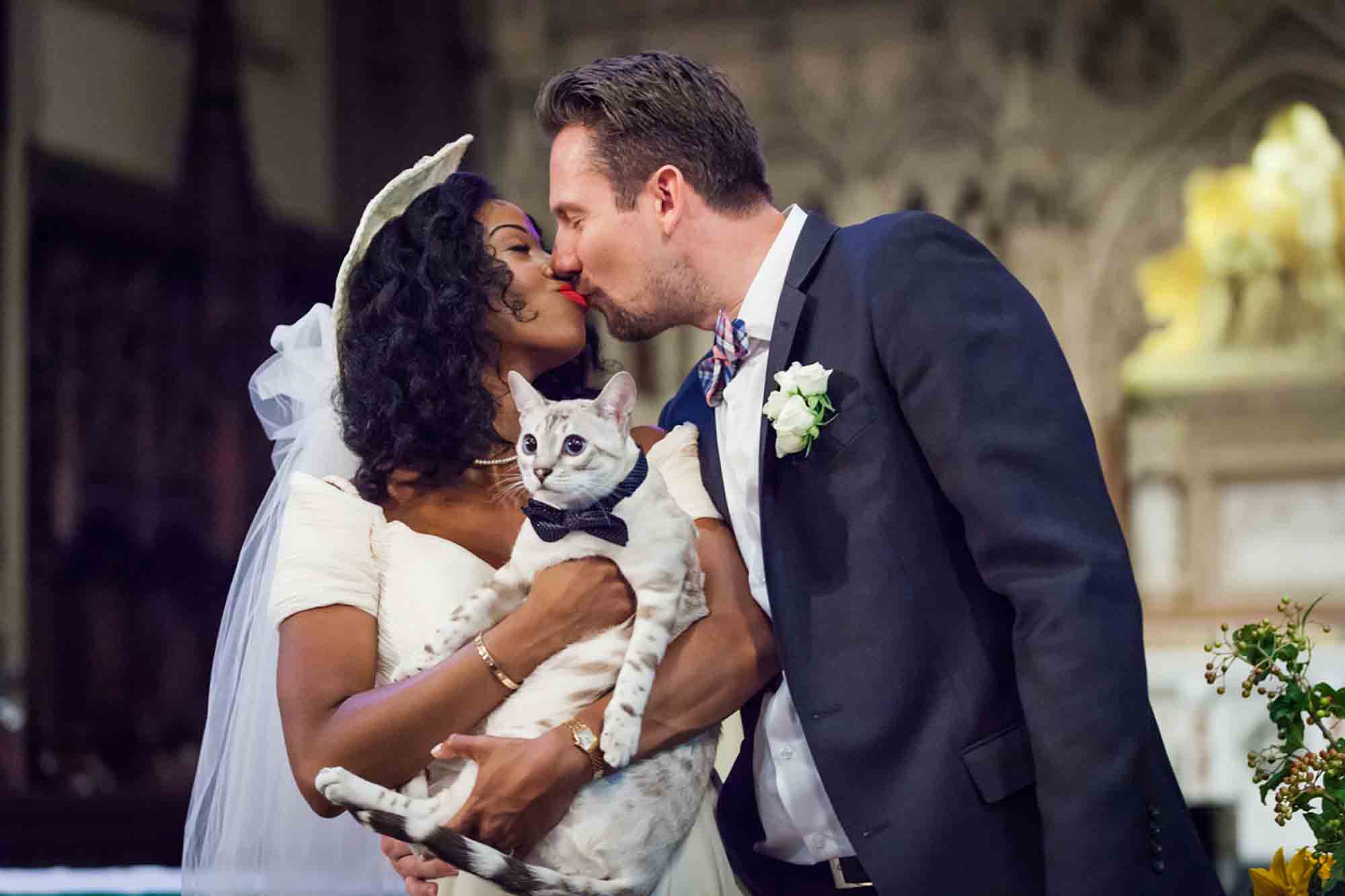 African American bride kissing groom at church while holding cat wearing black bowtie for an article on unexpected wedding photo tips