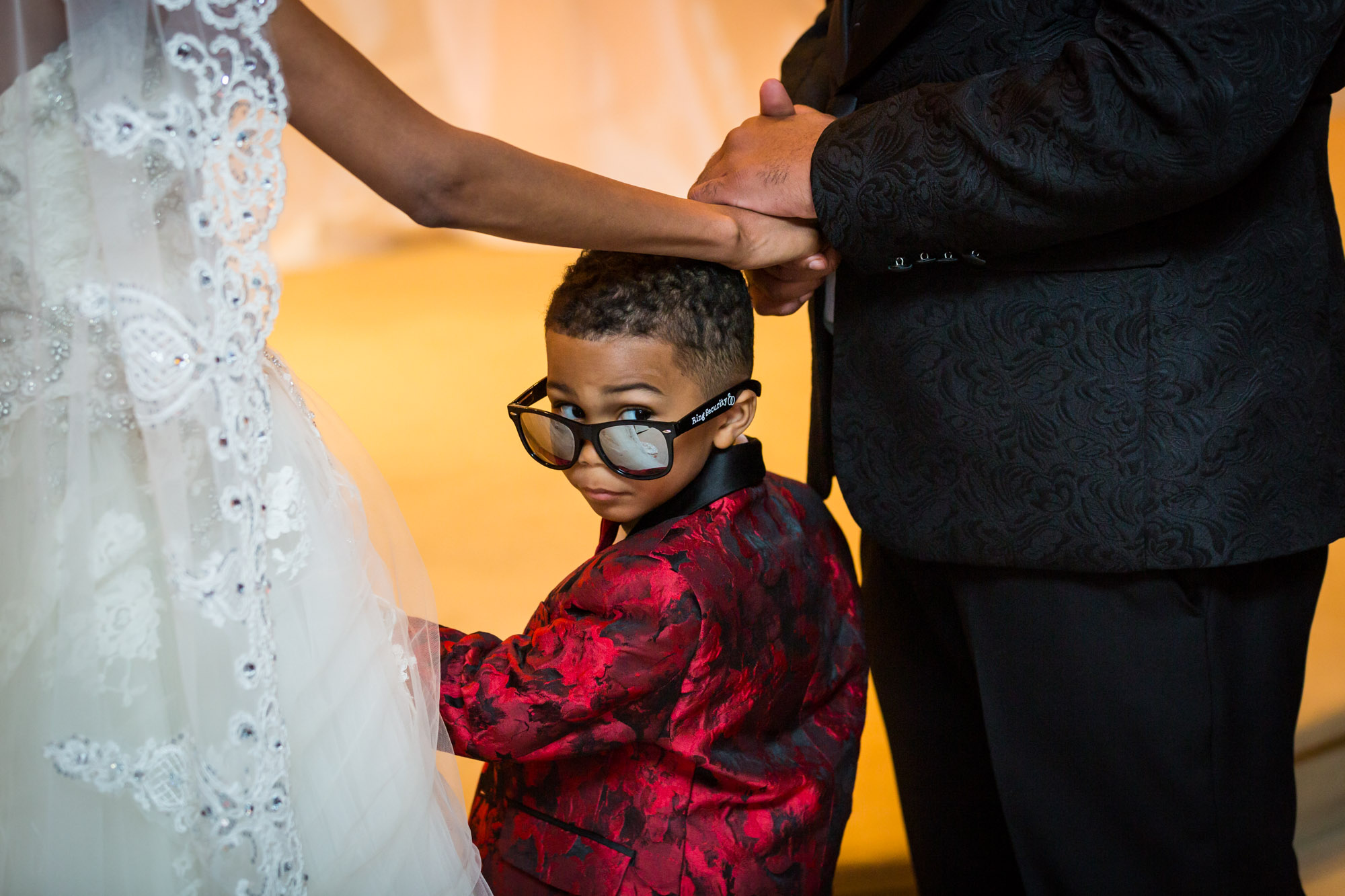 African American boy wearing red blazer and sun glasses looking over shoulder between bride and groom holding hands for an article on unexpected wedding photo tips