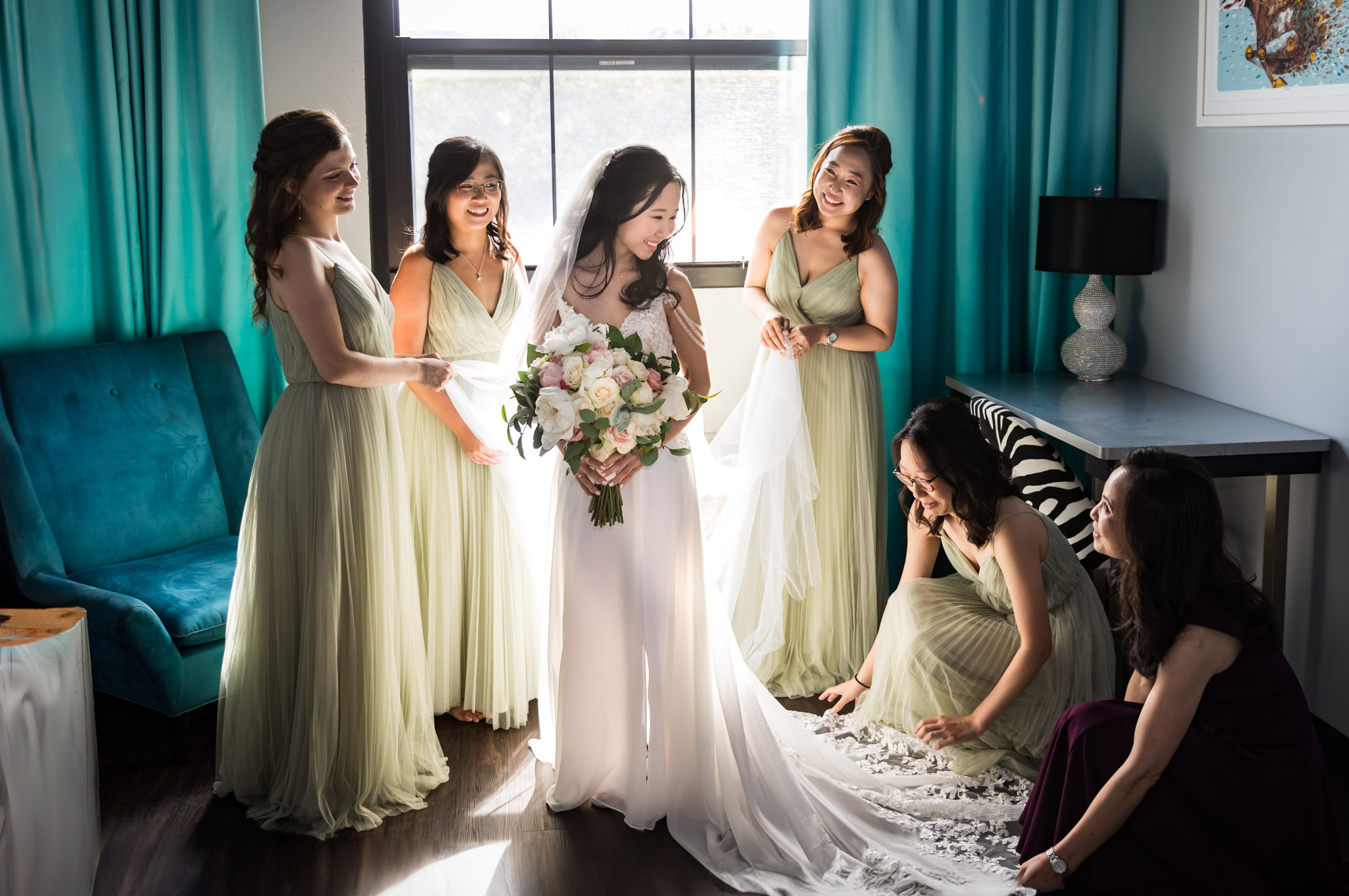 Bride holding large flower bouquet surrounded by bridesmaids attending to dress in front of window for an article on unexpected wedding photo tips