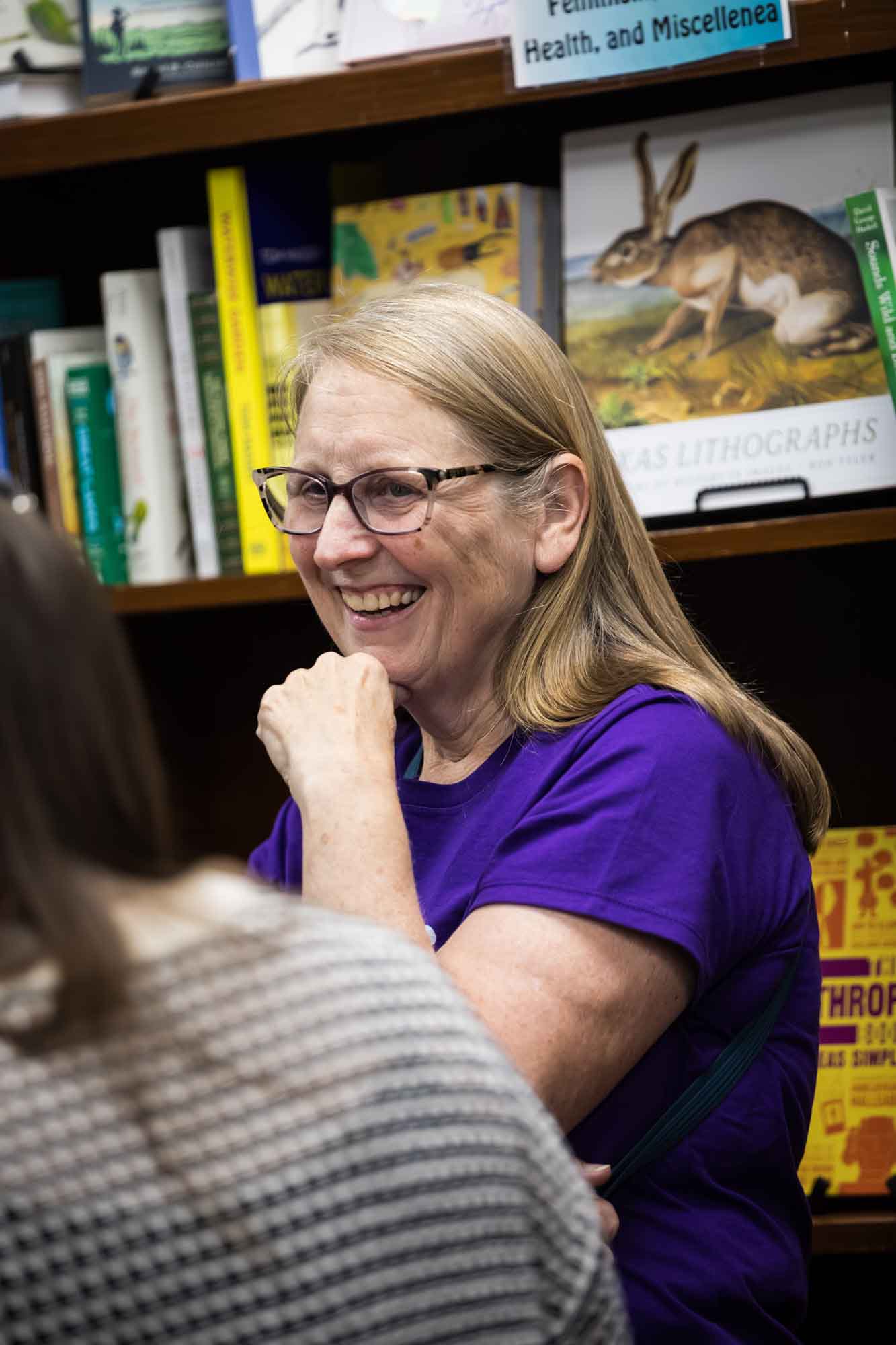 Austin book launch party photos of older woman smiling and wearing short-sleeved purple shirt in front of bookshelf