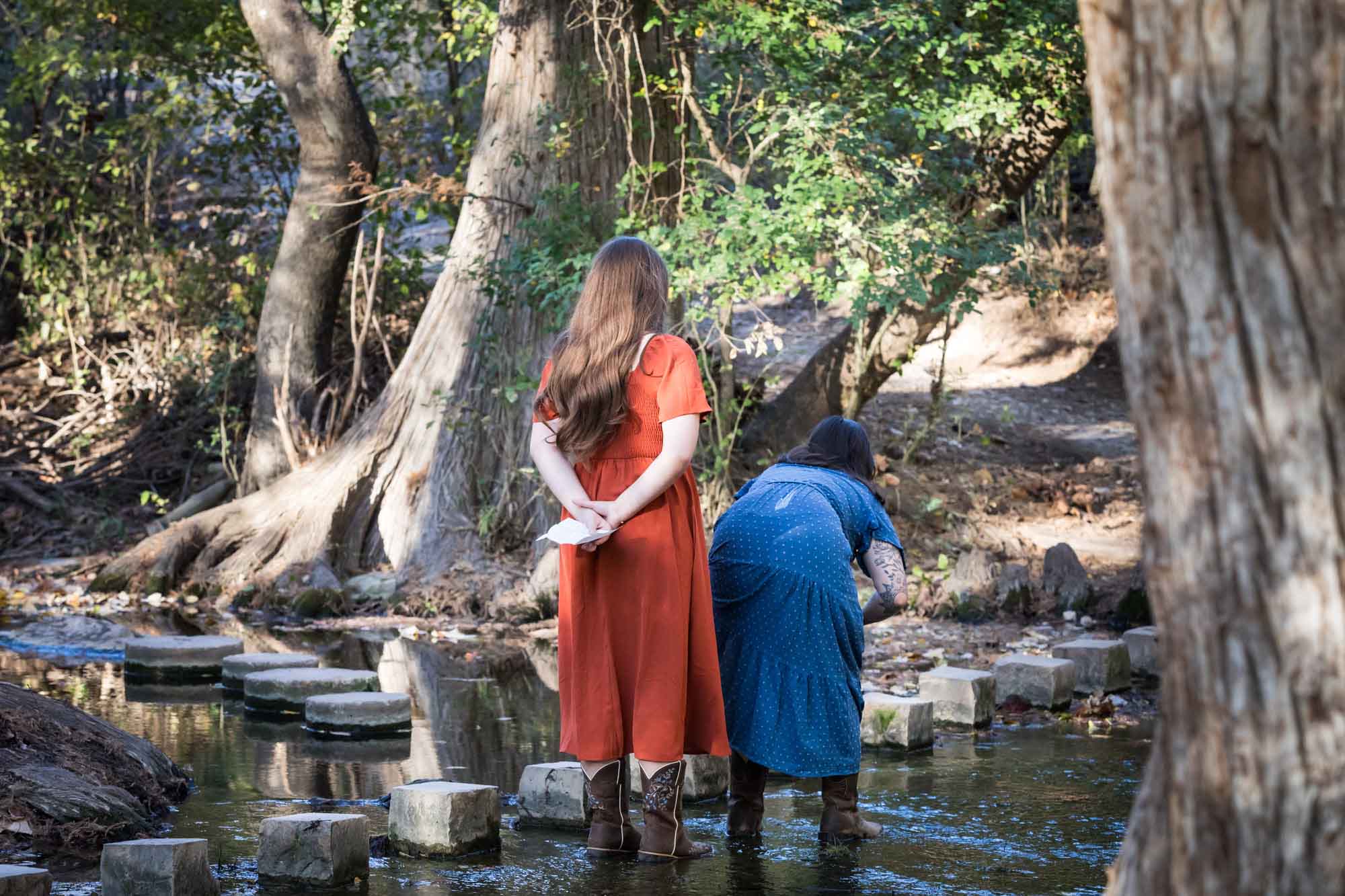Cibolo Nature Center surprise proposal photos of woman in orange dress holding piece of paper standing behind woman in blue dress in the forest