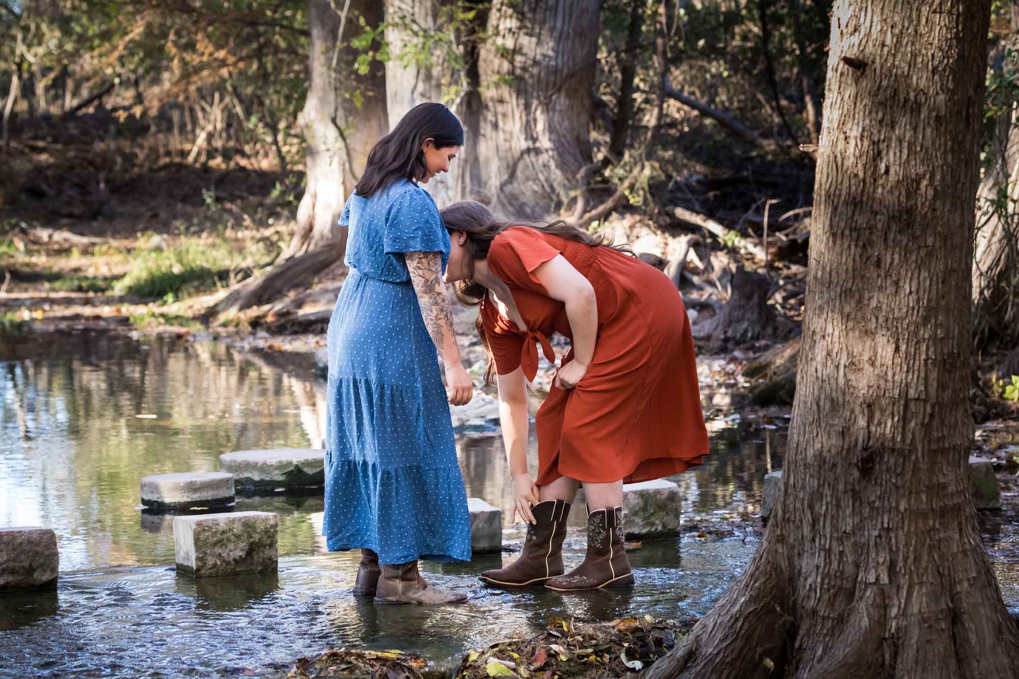 Cibolo Nature Center surprise proposal photos of woman in orange dress reaching down into boot standing in front of woman in blue dress in the forest