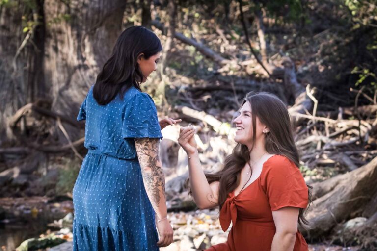Cibolo Nature Center surprise proposal photos of woman in orange dress on bended knee holding engagement ring in front of woman in blue dress in the forest