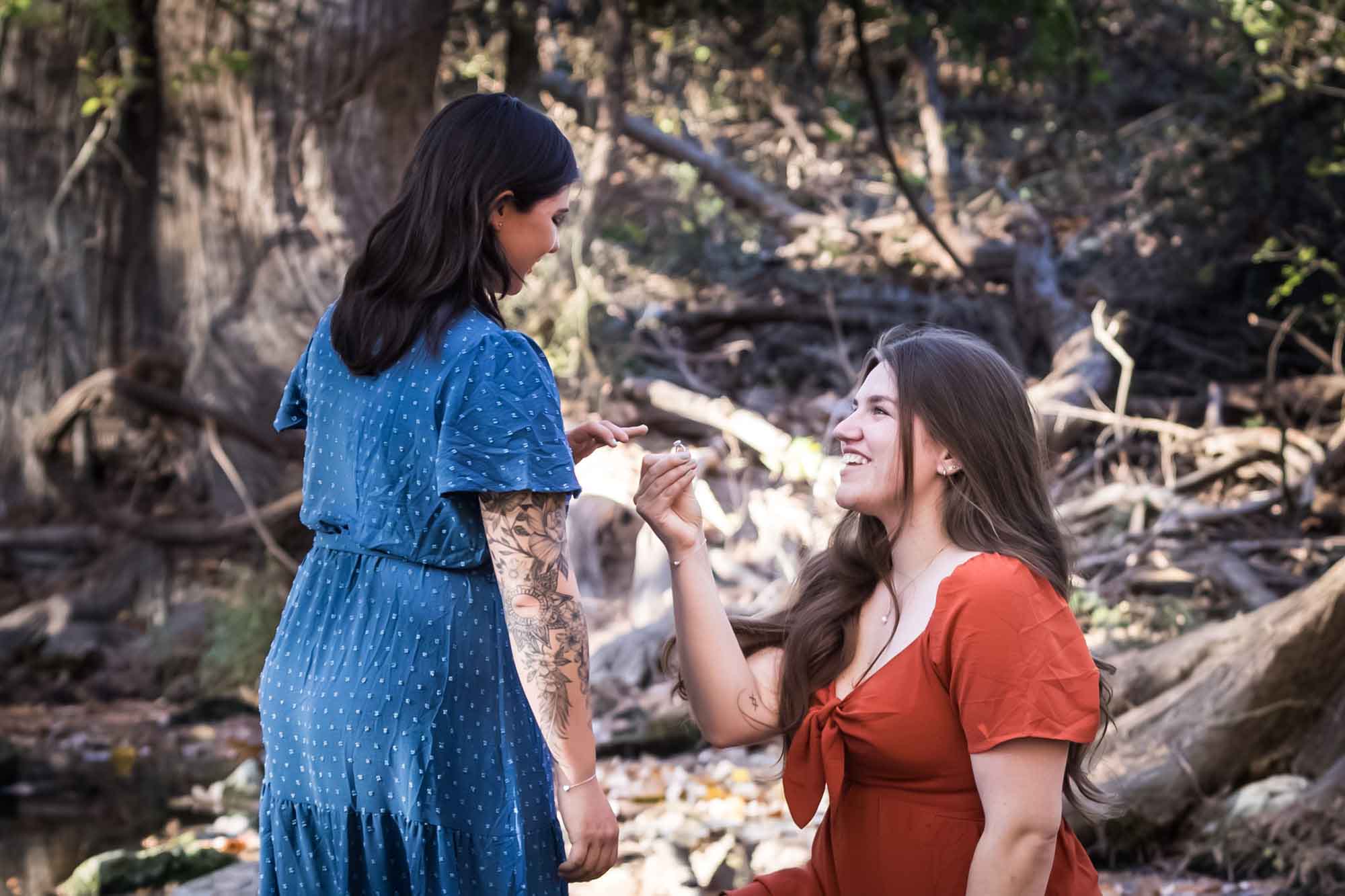 Cibolo Nature Center surprise proposal photos of woman in orange dress on bended knee holding engagement ring in front of woman in blue dress in the forest