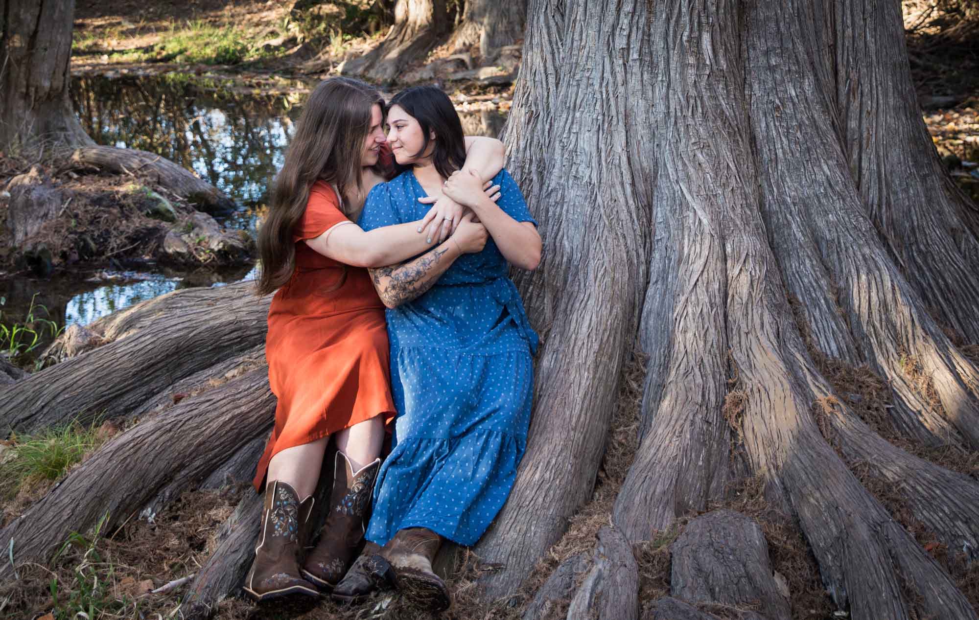 Cibolo Nature Center surprise proposal photos of woman in orange dress hugging woman in blue dress sitting on roots of large tree in the forest