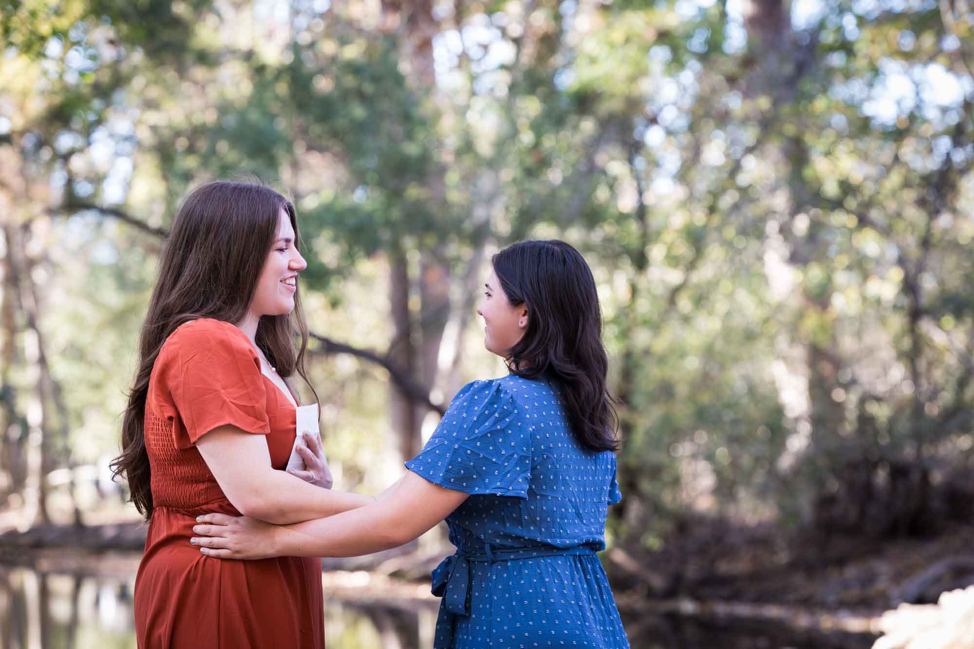Cibolo Nature Center surprise proposal photos of woman in orange dress holding piece of paper standing in front of woman in blue dress in the forest