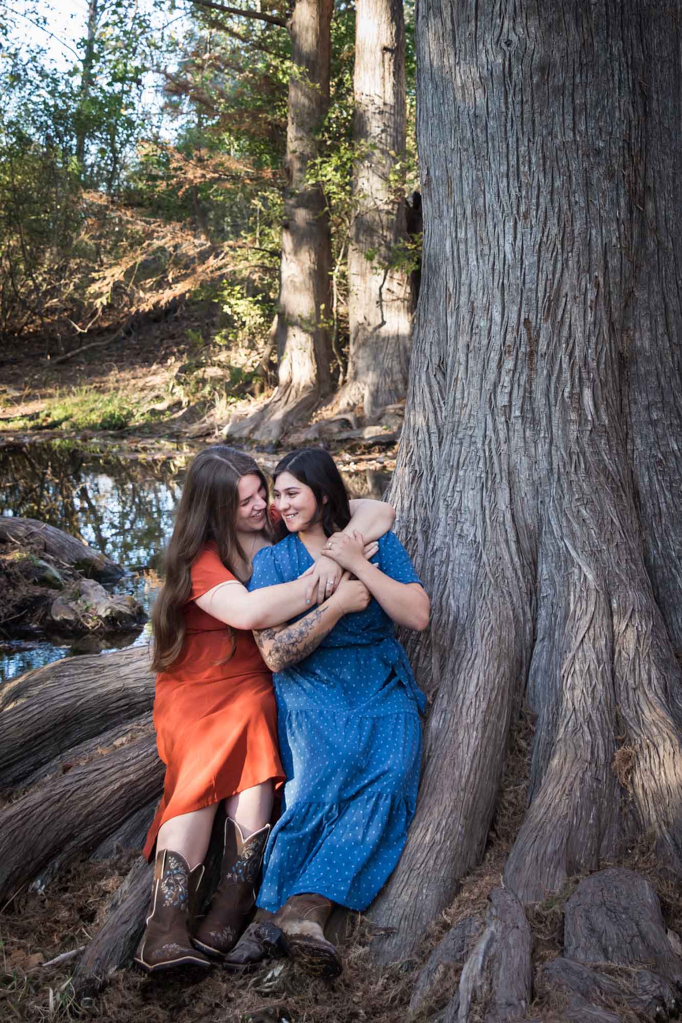 Cibolo Nature Center surprise proposal photos of woman in orange dress hugging woman in blue dress sitting on roots of large tree in the forest