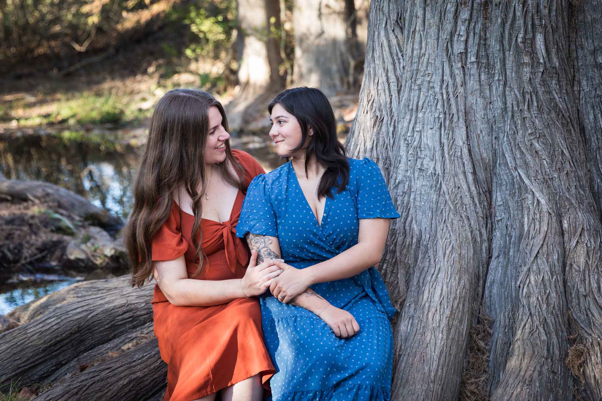 Cibolo Nature Center surprise proposal photos of woman in orange dress hugging woman in blue dress sitting on roots of large tree in the forest
