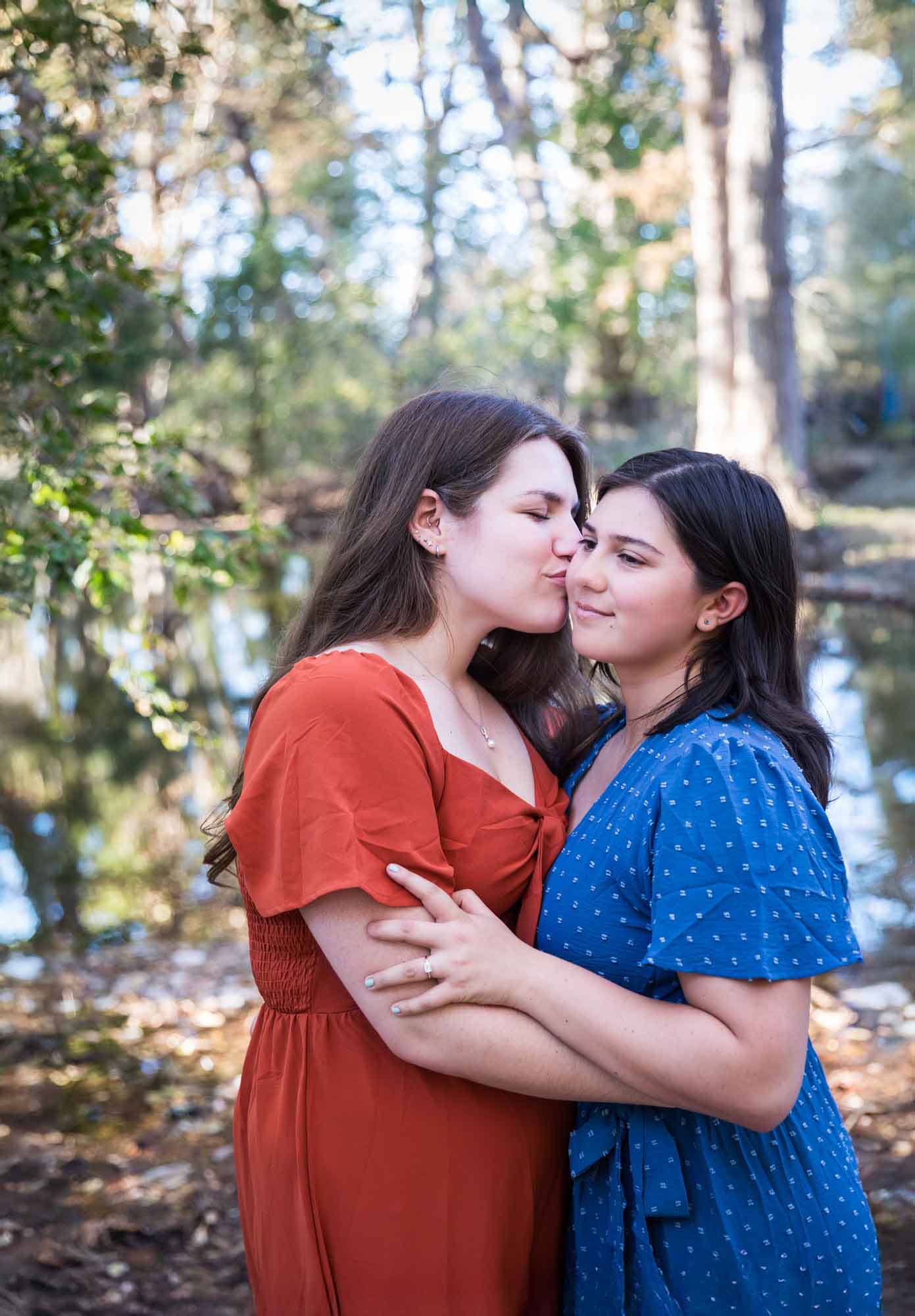 Cibolo Nature Center surprise proposal photos of woman in orange dress kissing cheek of woman in blue dress in the forest