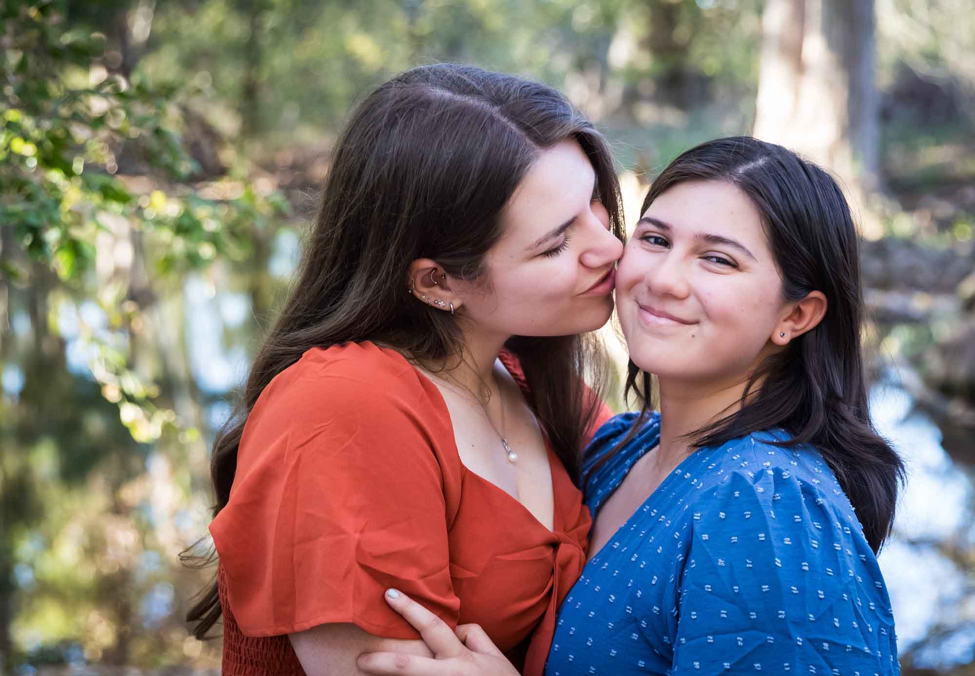 Cibolo Nature Center surprise proposal photos of woman in orange dress kissing cheek of woman in blue dress in the forest