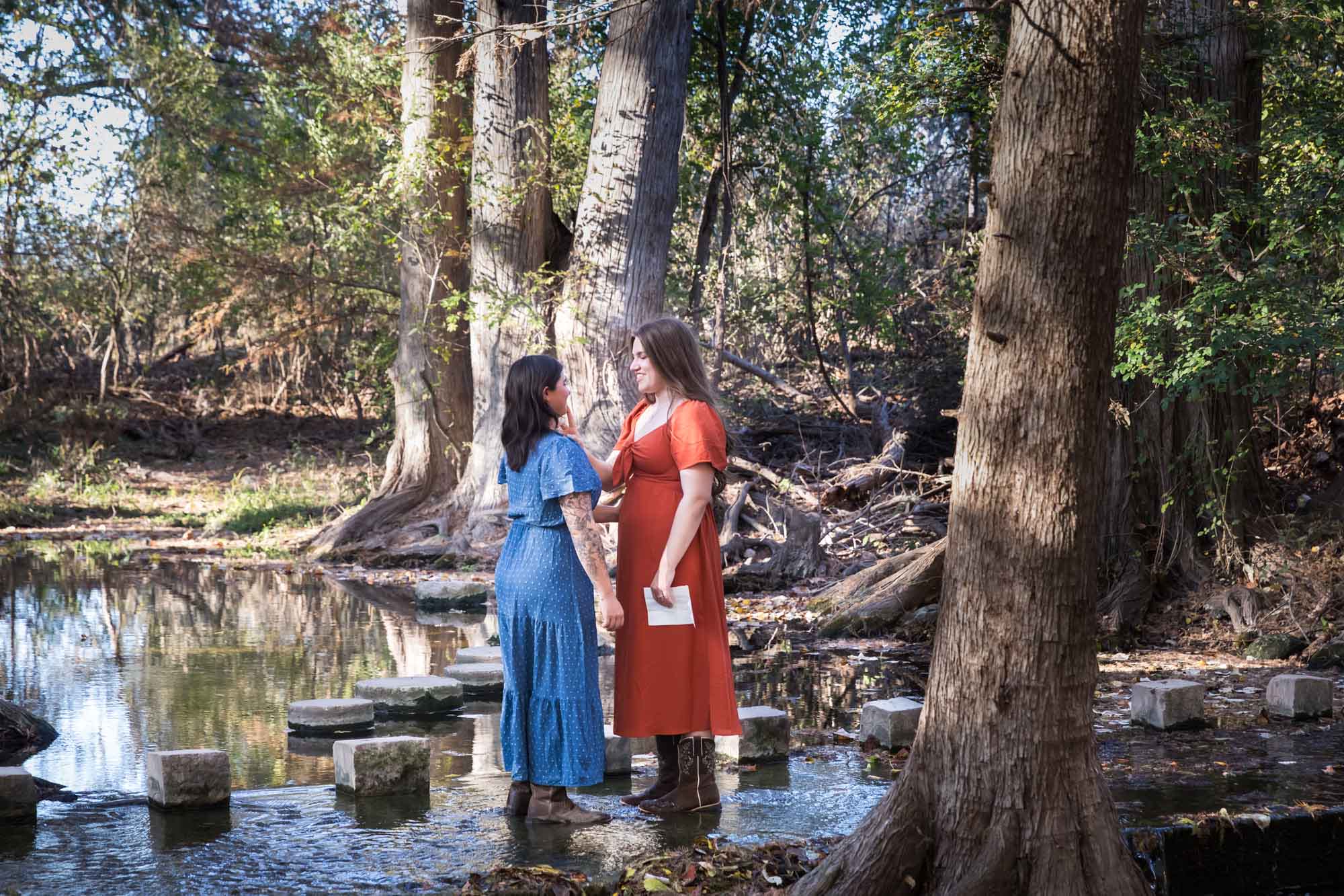 Cibolo Nature Center surprise proposal photos of woman in orange dress holding piece of paper standing in front of woman in blue dress in the forest