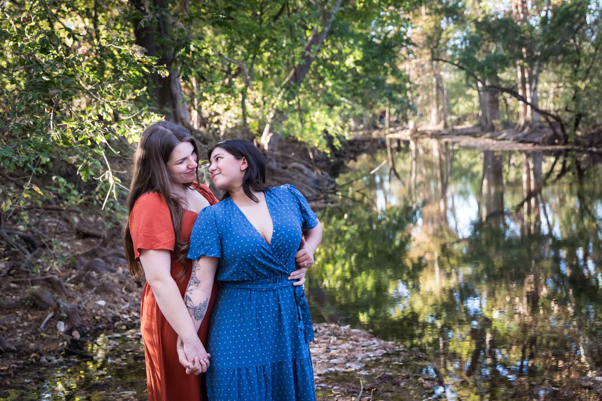 Cibolo Nature Center surprise proposal photos of woman in orange dress and woman in blue dress holding hands in the forest with Cibolo Creek in background