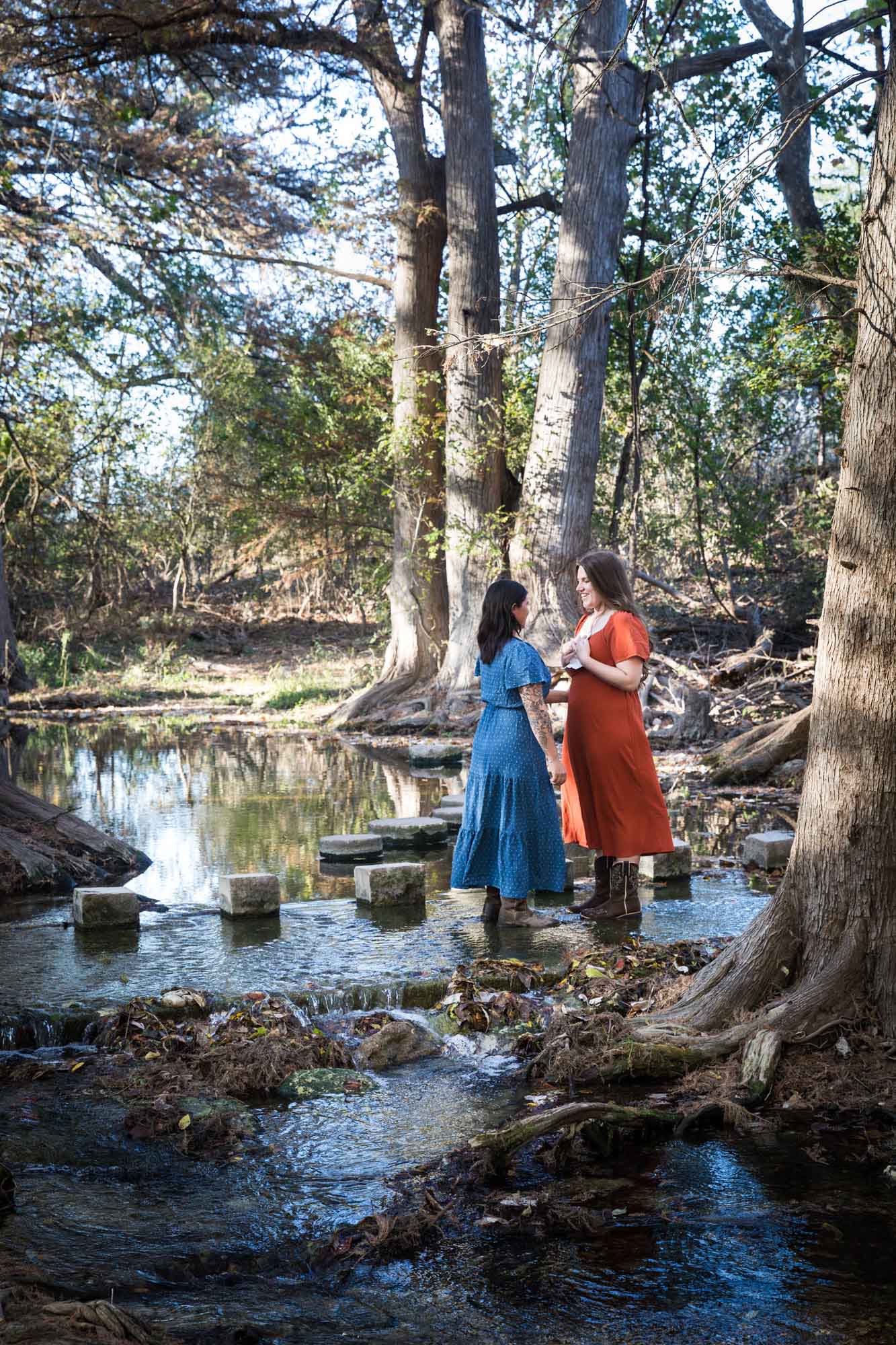 Cibolo Nature Center surprise proposal photos of woman in orange dress holding piece of paper standing in front of woman in blue dress in the forest