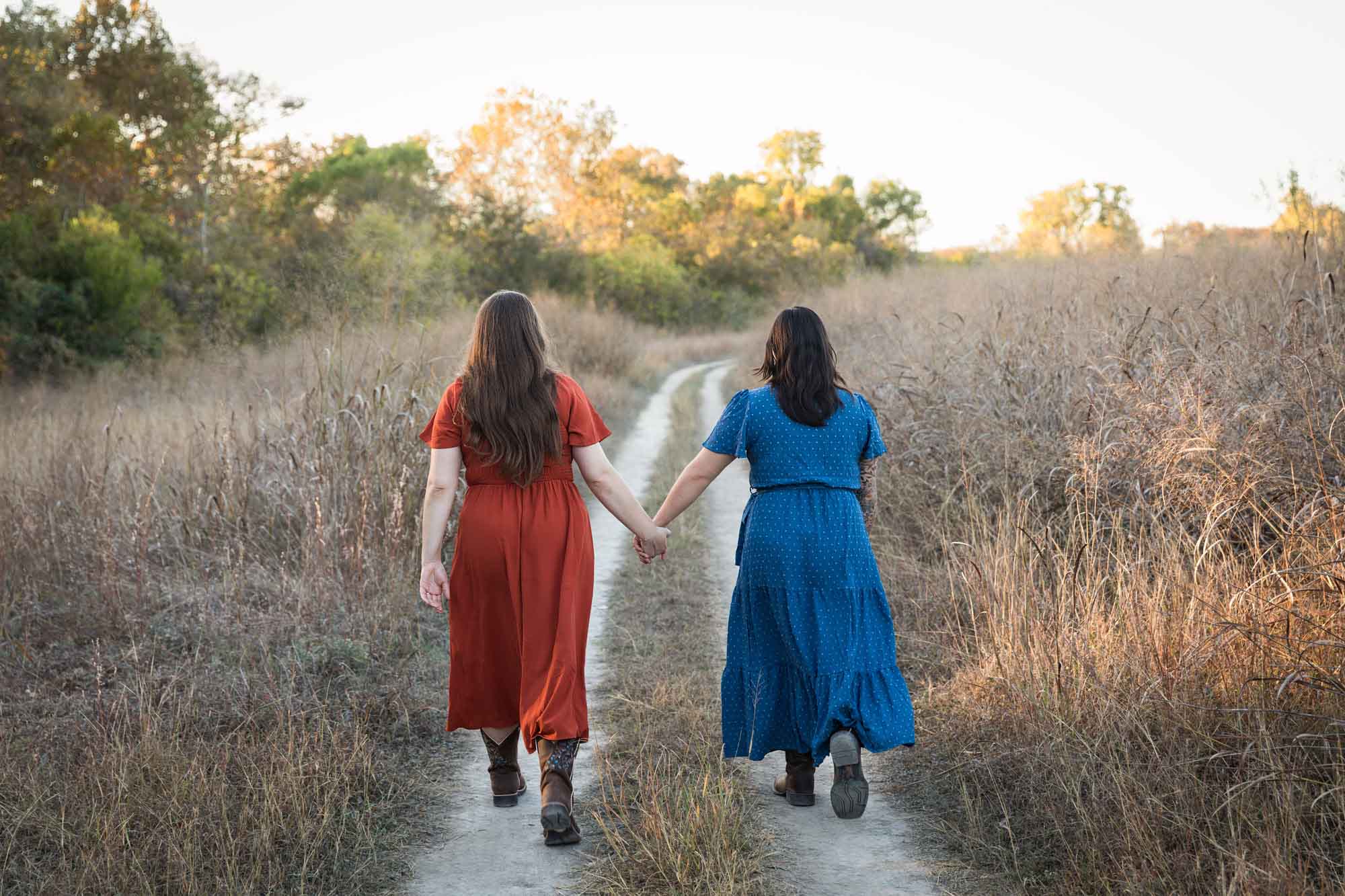 Cibolo Nature Center surprise proposal photos of woman in orange dress holding hands and walking with woman in blue dress down a dirt road