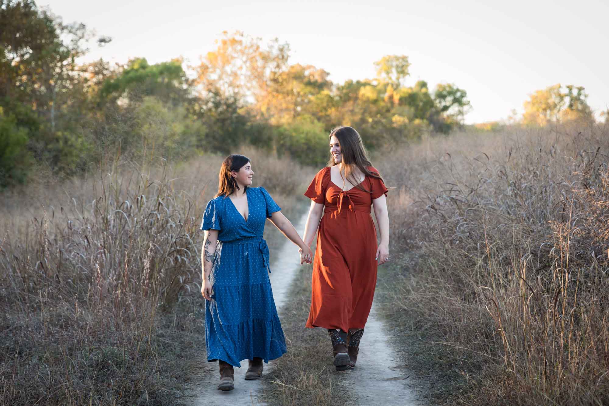 Cibolo Nature Center surprise proposal photos of woman in orange dress holding hands and walking with woman in blue dress down a dirt road
