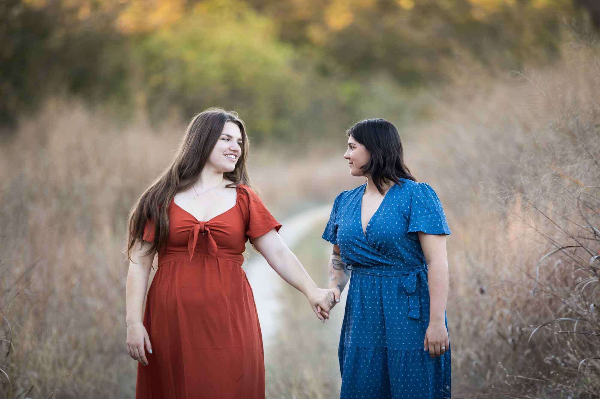 Cibolo Nature Center surprise proposal photos of woman in orange dress holding hands and walking with woman in blue dress down a dirt road