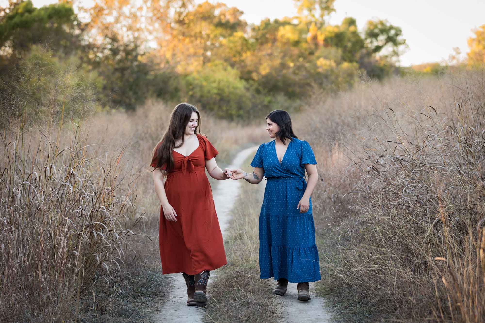 Cibolo Nature Center surprise proposal photos of woman in orange dress holding hands and walking with woman in blue dress down a dirt road