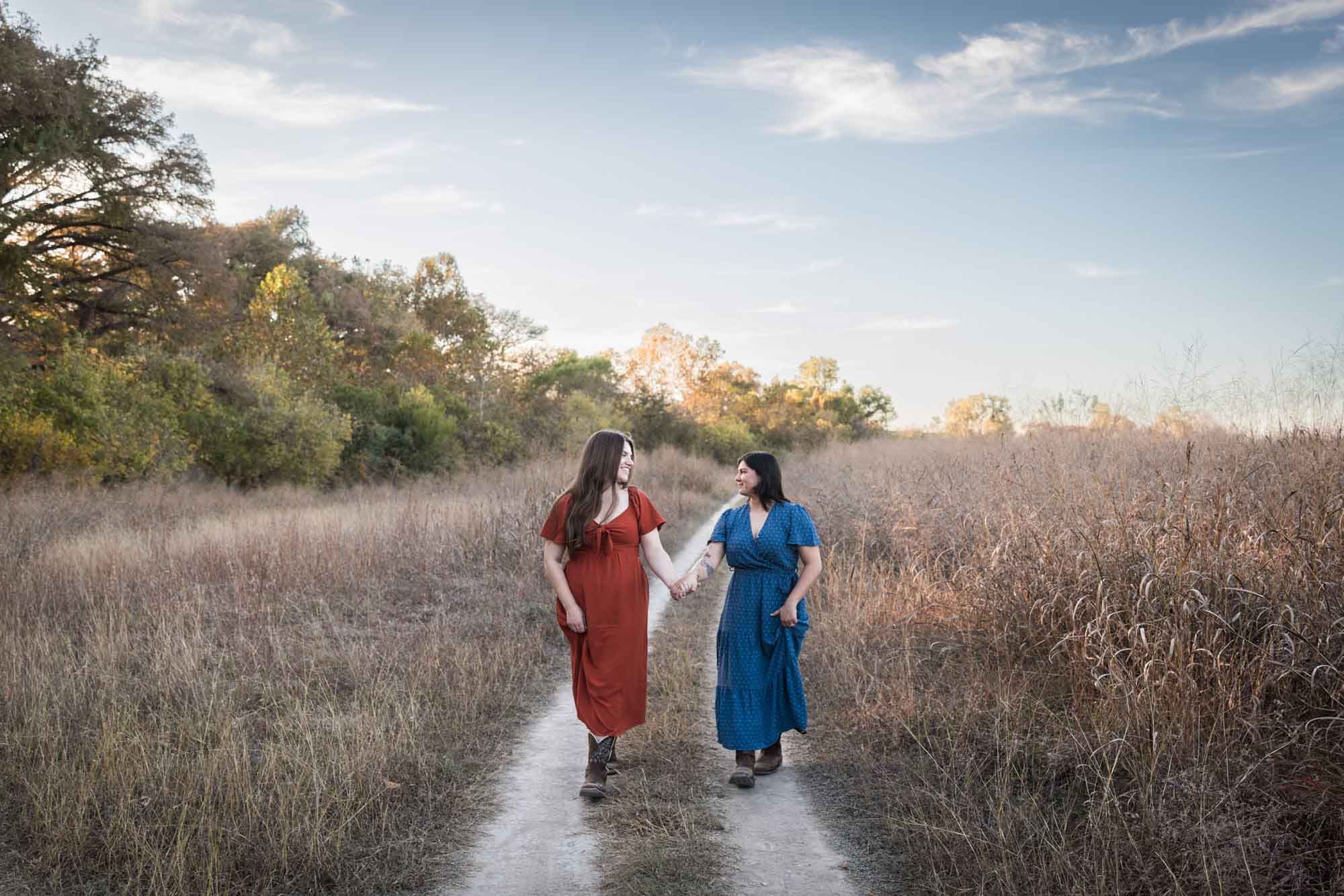 Cibolo Nature Center surprise proposal photos of woman in orange dress holding hands and walking with woman in blue dress down a dirt road