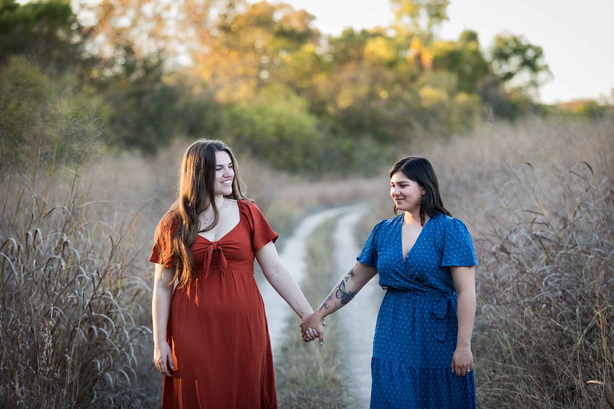 Cibolo Nature Center surprise proposal photos of woman in orange dress holding hands and walking with woman in blue dress down a dirt road