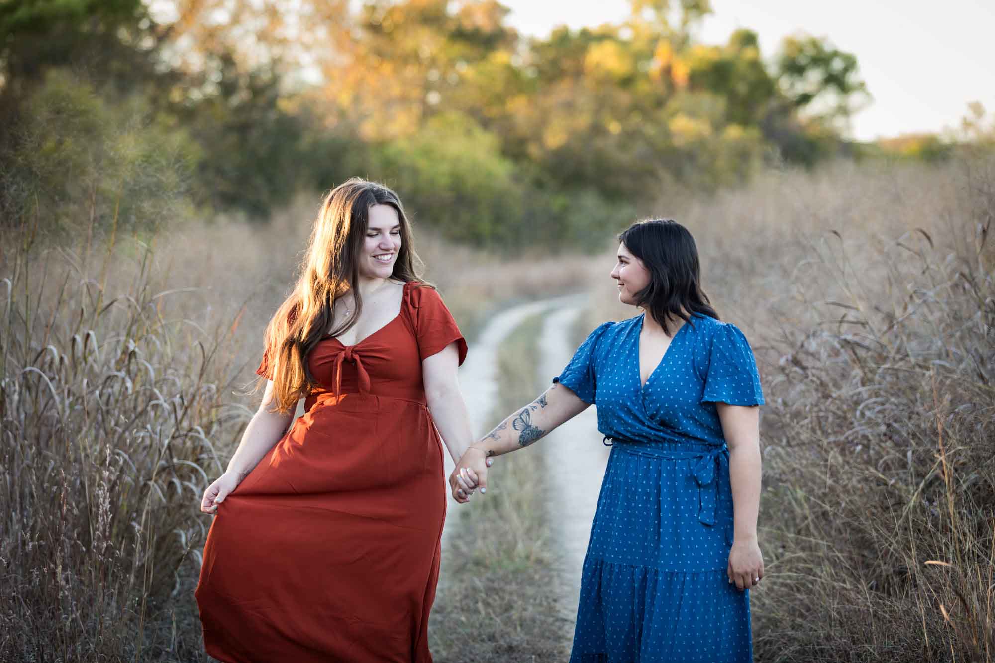 Cibolo Nature Center surprise proposal photos of woman in orange dress holding hands and walking with woman in blue dress down a dirt road