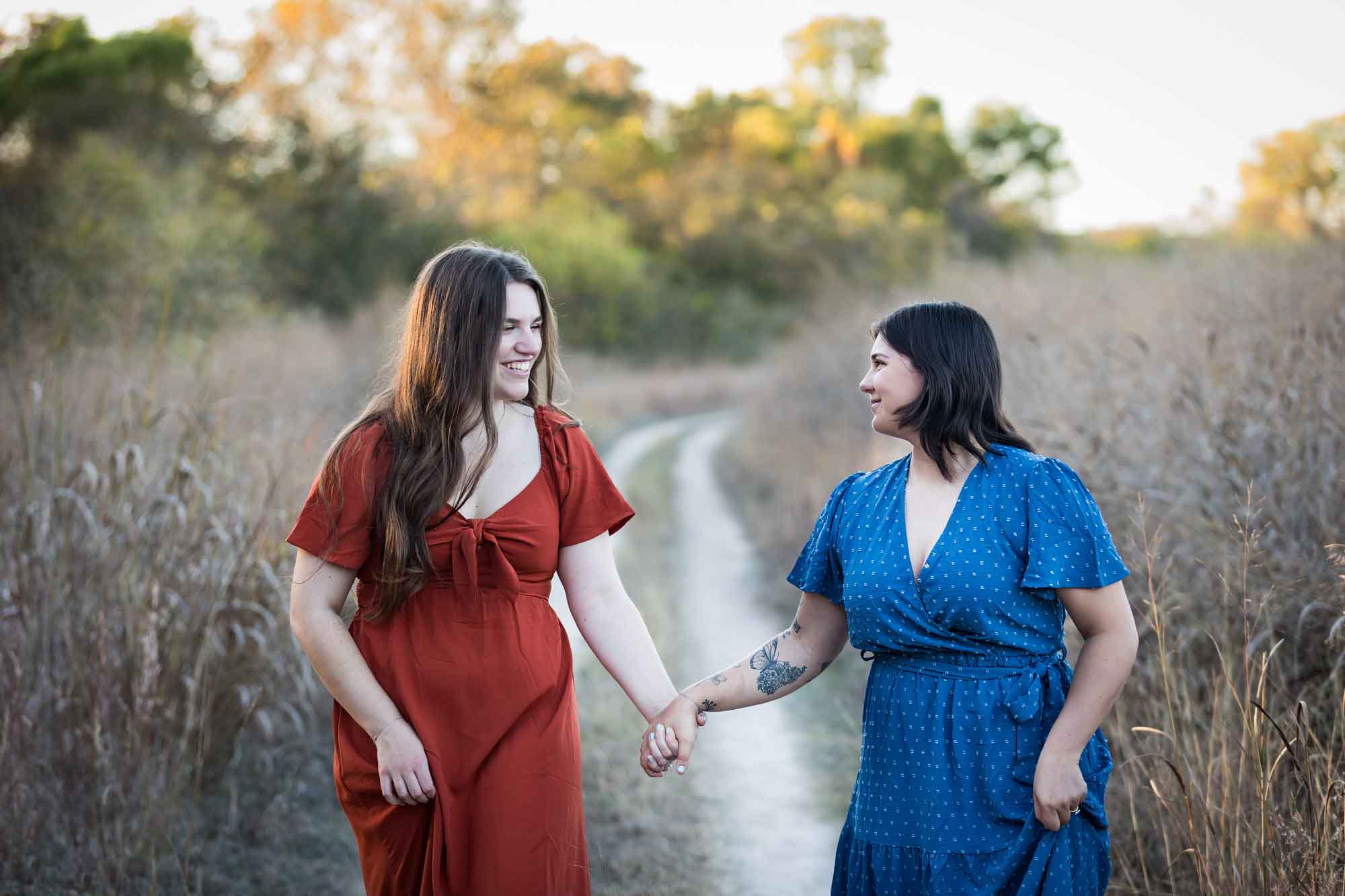 Cibolo Nature Center surprise proposal photos of woman in orange dress holding hands and walking with woman in blue dress down a dirt road