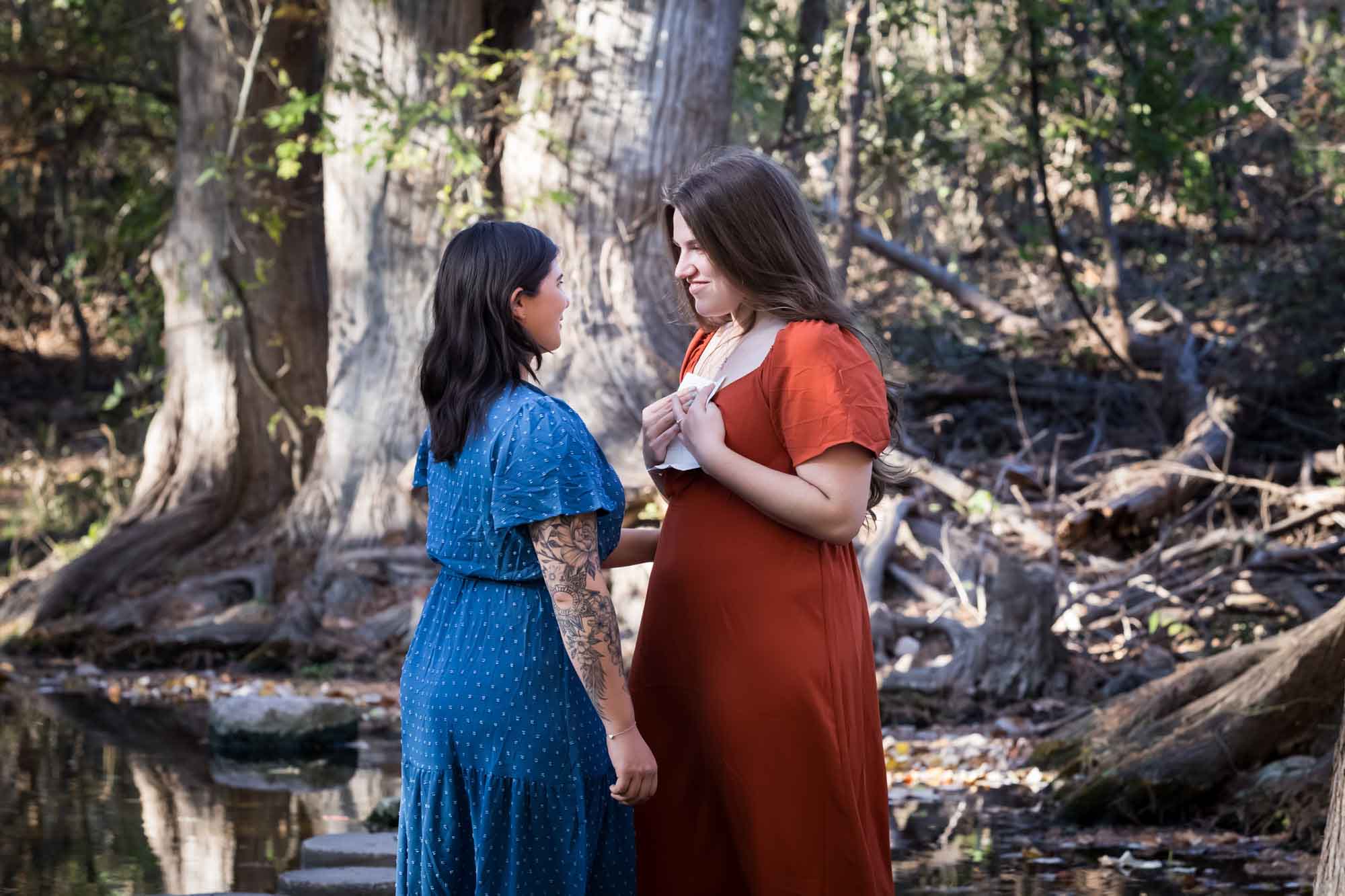Cibolo Nature Center surprise proposal photos of woman in orange dress holding piece of paper standing in front of woman in blue dress in the forest