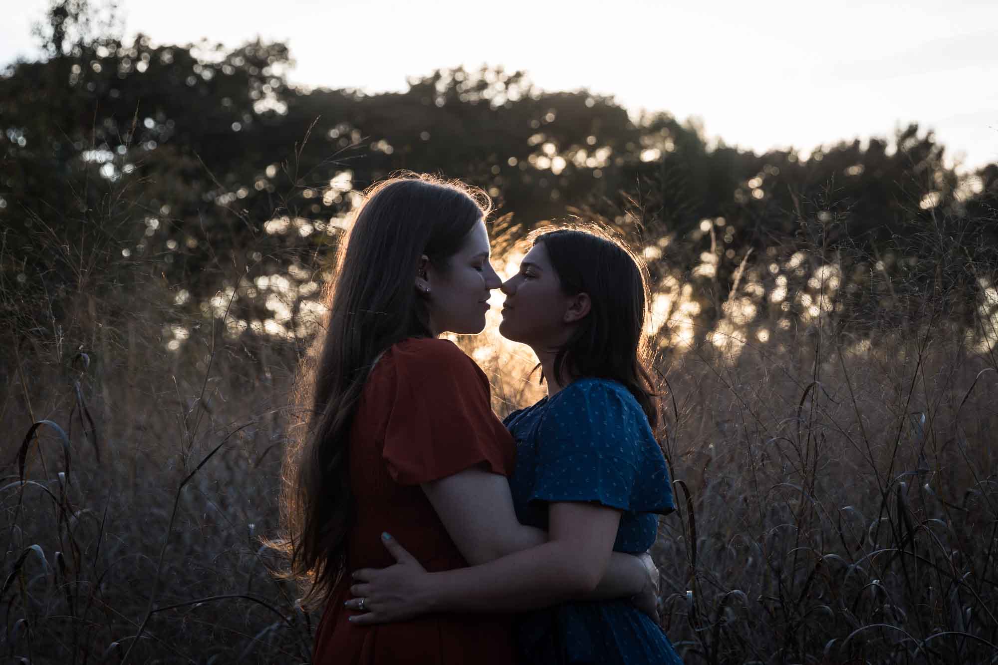 Silhouette photo of woman in orange dress kissing woman in blue dress in the middle of marsh grass at sunset