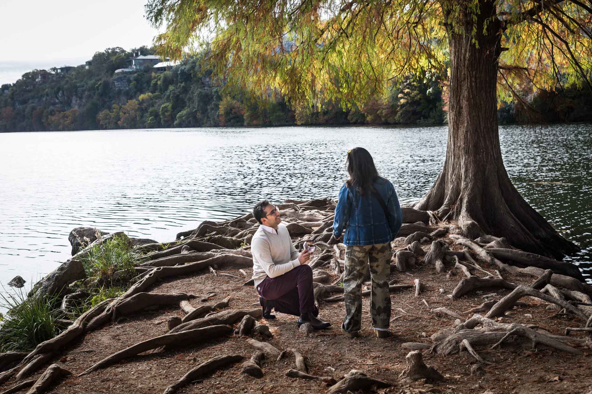 Man wearing maroon pants and white shirt standing on bended knee holding ring box in front of woman wearing camo pants and denim jacket at end of island with tree in background during a Red Bud Isle surprise proposal