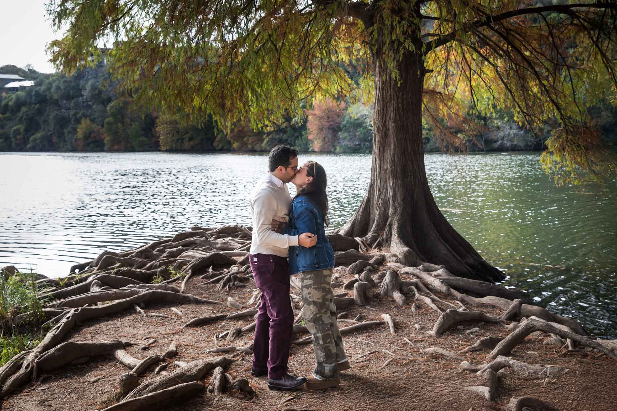 Man wearing maroon pants and white shirt kissing woman wearing camo pants and denim jacket at end of island with tree in background during a Red Bud Isle surprise proposal