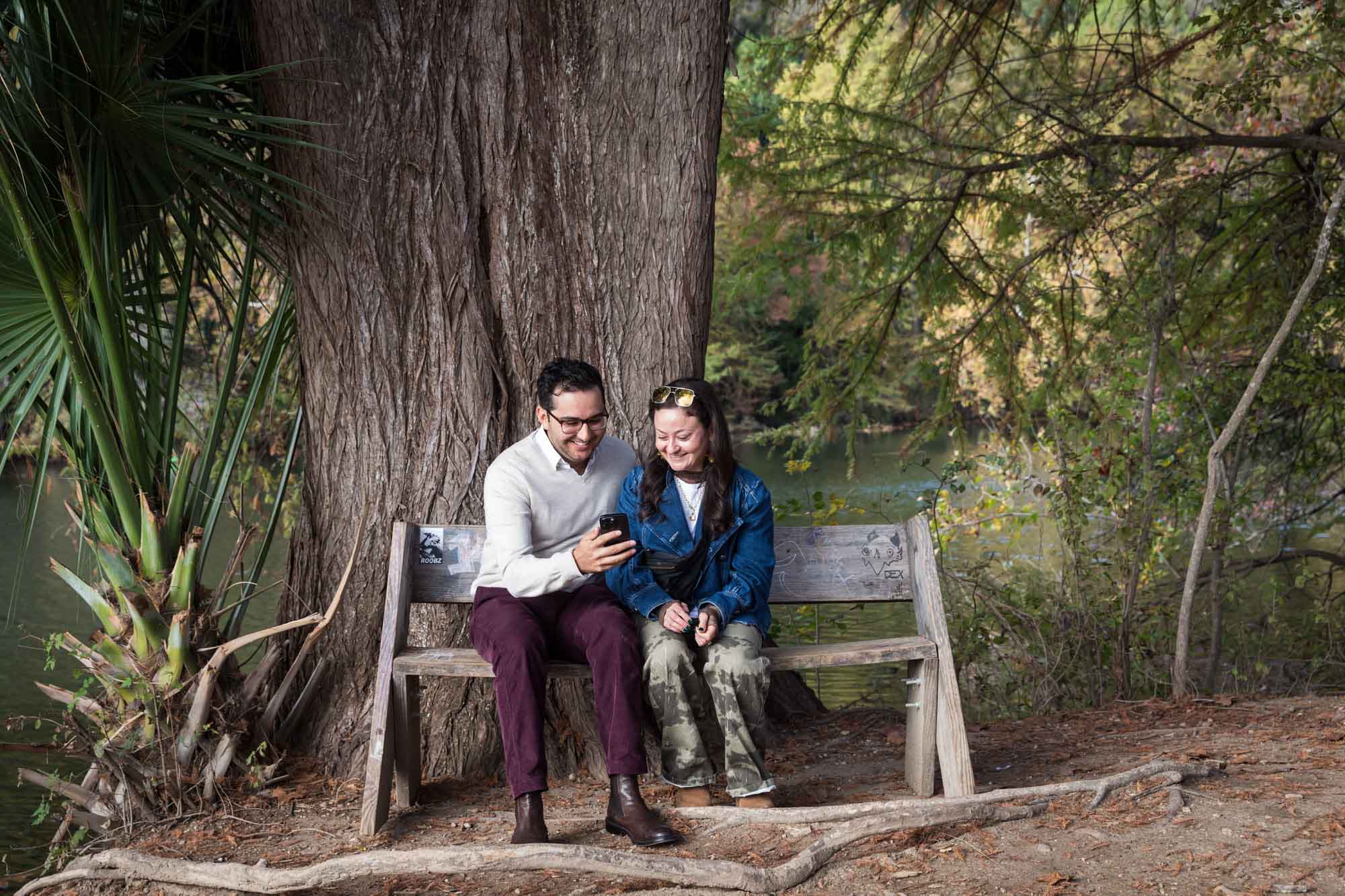 Man wearing maroon pants and grey sweater sitting on bench beside woman wearing camo pants and denim jacket in front of tree during a Red Bud Isle surprise proposal