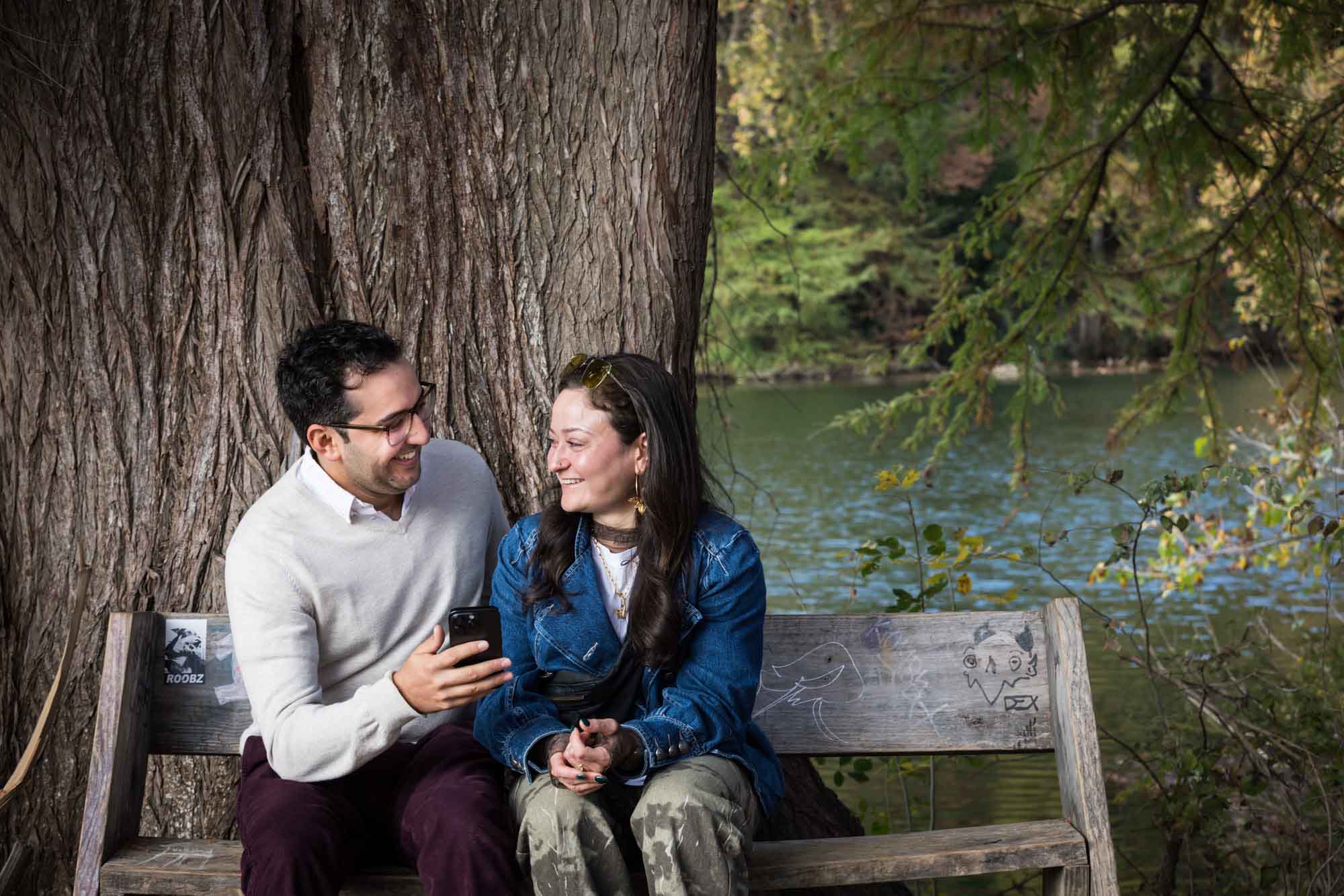 Man wearing maroon pants and grey sweater sitting on bench beside woman wearing camo pants and denim jacket in front of tree during a Red Bud Isle surprise proposal