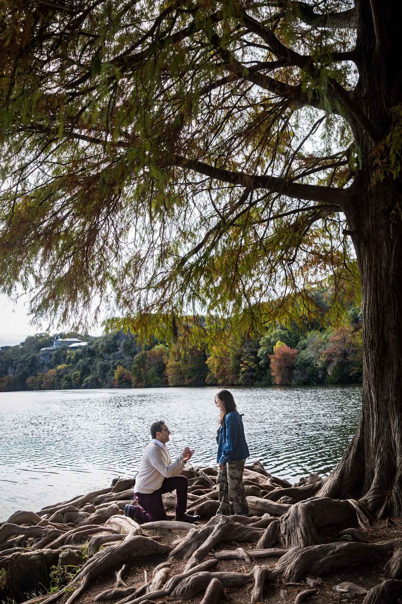 Man wearing maroon pants and white shirt standing on bended knee holding ring box in front of woman wearing camo pants and denim jacket at end of island with tree in background during a Red Bud Isle surprise proposal