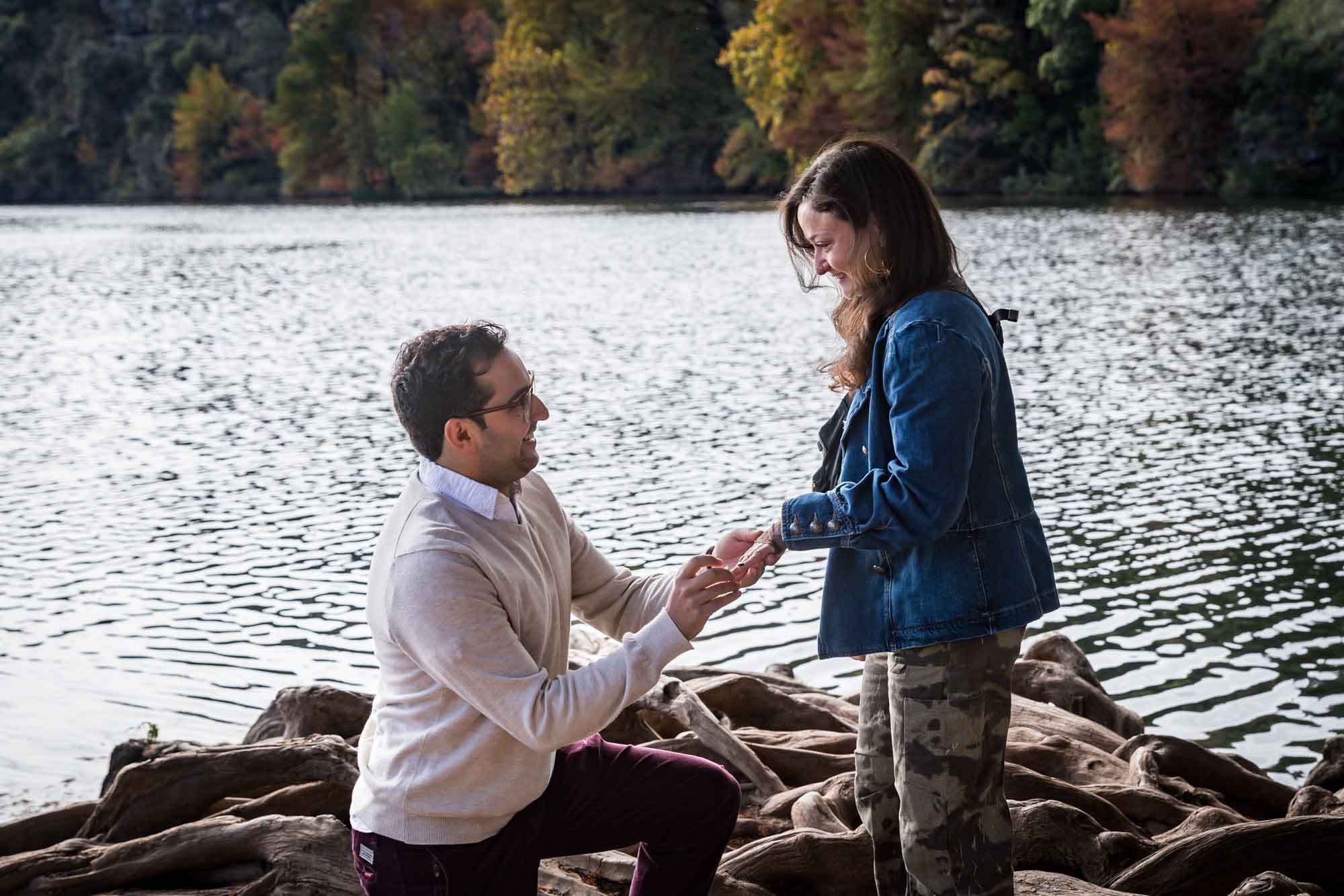 Man wearing maroon pants and white shirt standing on bended knee putting ring on finger of woman wearing camo pants and denim jacket at end of island with tree in background during a Red Bud Isle surprise proposal