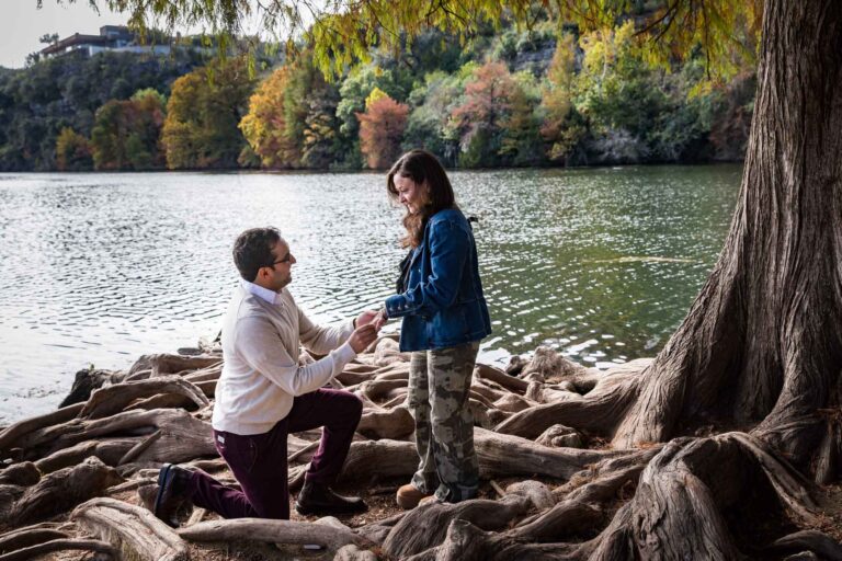 Man wearing maroon pants and white shirt standing on bended knee putting ring on finger of woman wearing camo pants and denim jacket at end of island with tree in background during a Red Bud Isle surprise proposal