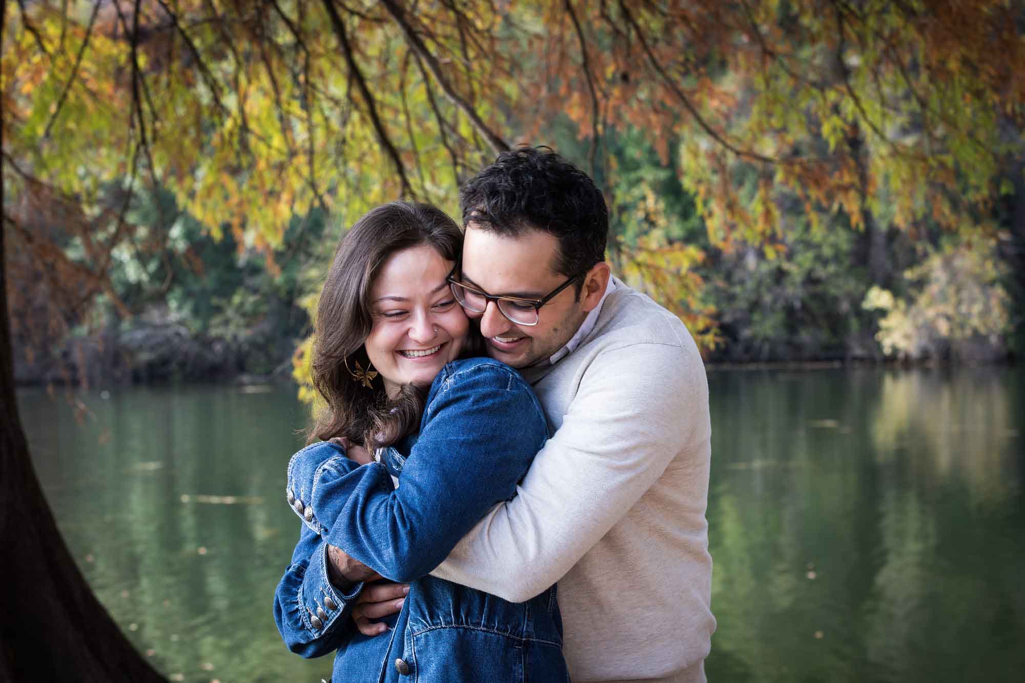 Man wearing grey sweater hugging woman wearing denim jacket in front of river and tree with colorful leaves during a Red Bud Isle surprise proposal and engagement portrait
