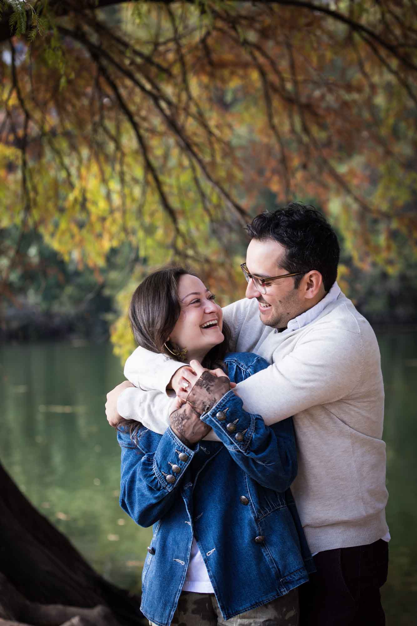 Man wearing grey sweater hugging woman wearing denim jacket in front of river and tree with colorful leaves during a Red Bud Isle surprise proposal and engagement portrait