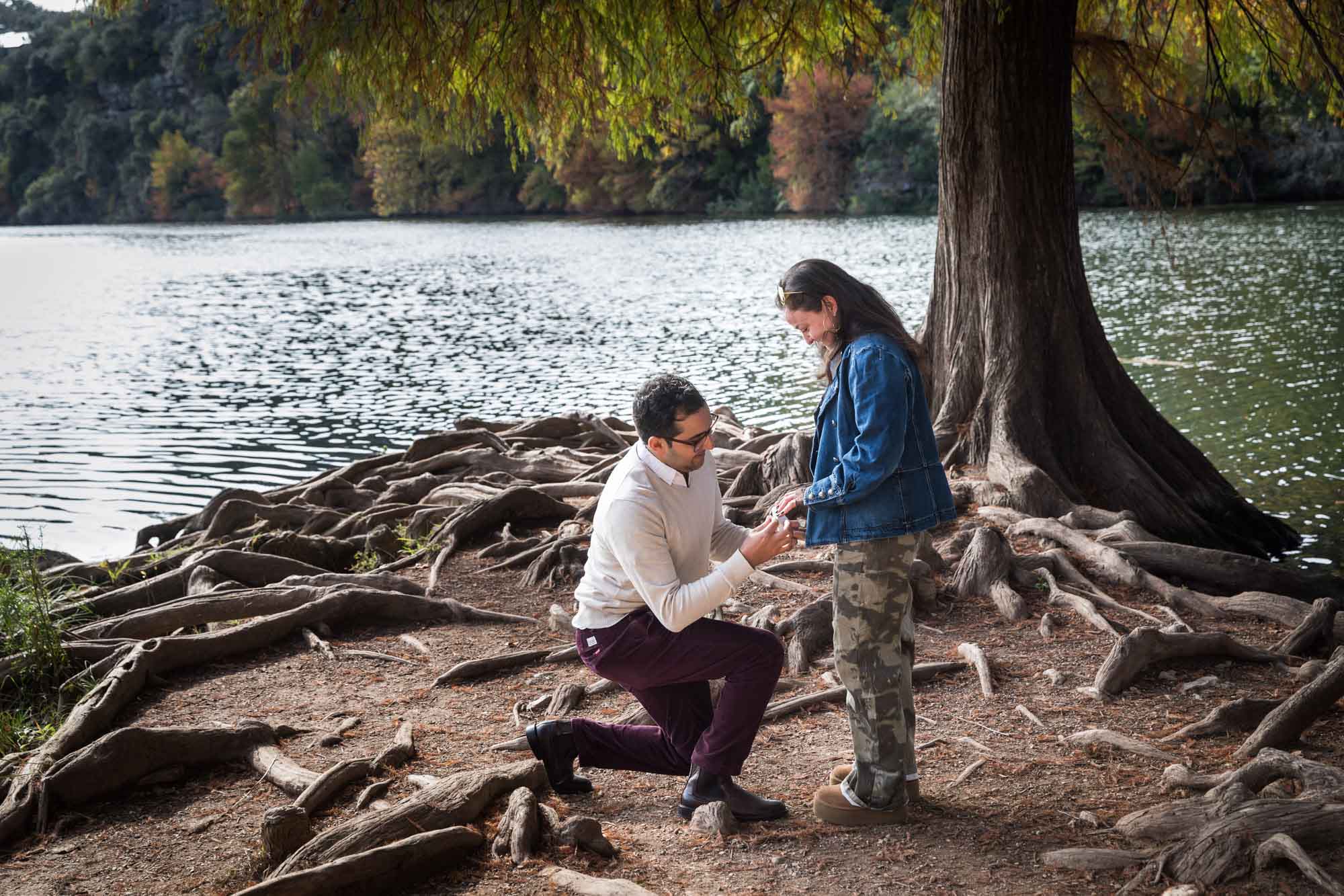 Man wearing maroon pants and white shirt standing on bended knee putting ring on finger of woman wearing camo pants and denim jacket at end of island with tree in background during a Red Bud Isle surprise proposal