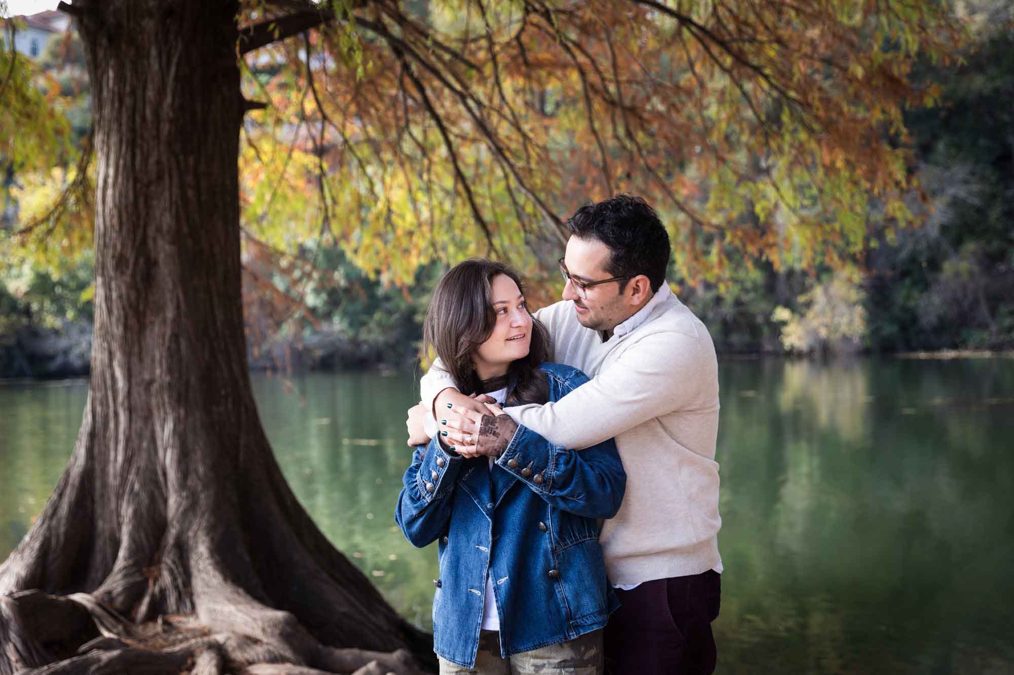 Man wearing grey sweater hugging woman wearing denim jacket in front of river and tree with colorful leaves during a Red Bud Isle surprise proposal and engagement portrait